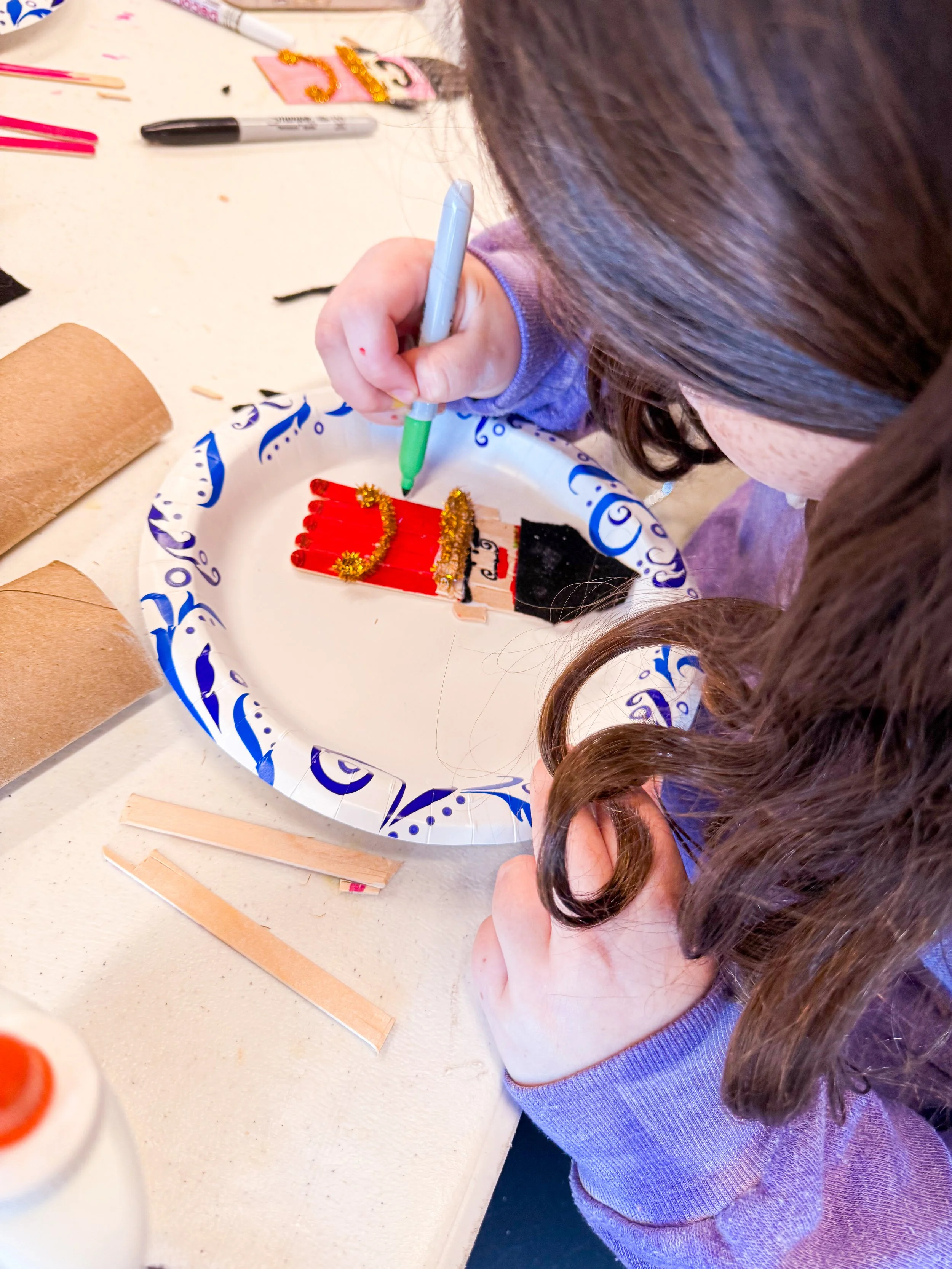 A child decorating a small popsicle stick Christmas craft with toothpicks and gold tinsel on a paper plate, surrounded by craft supplies on a table.