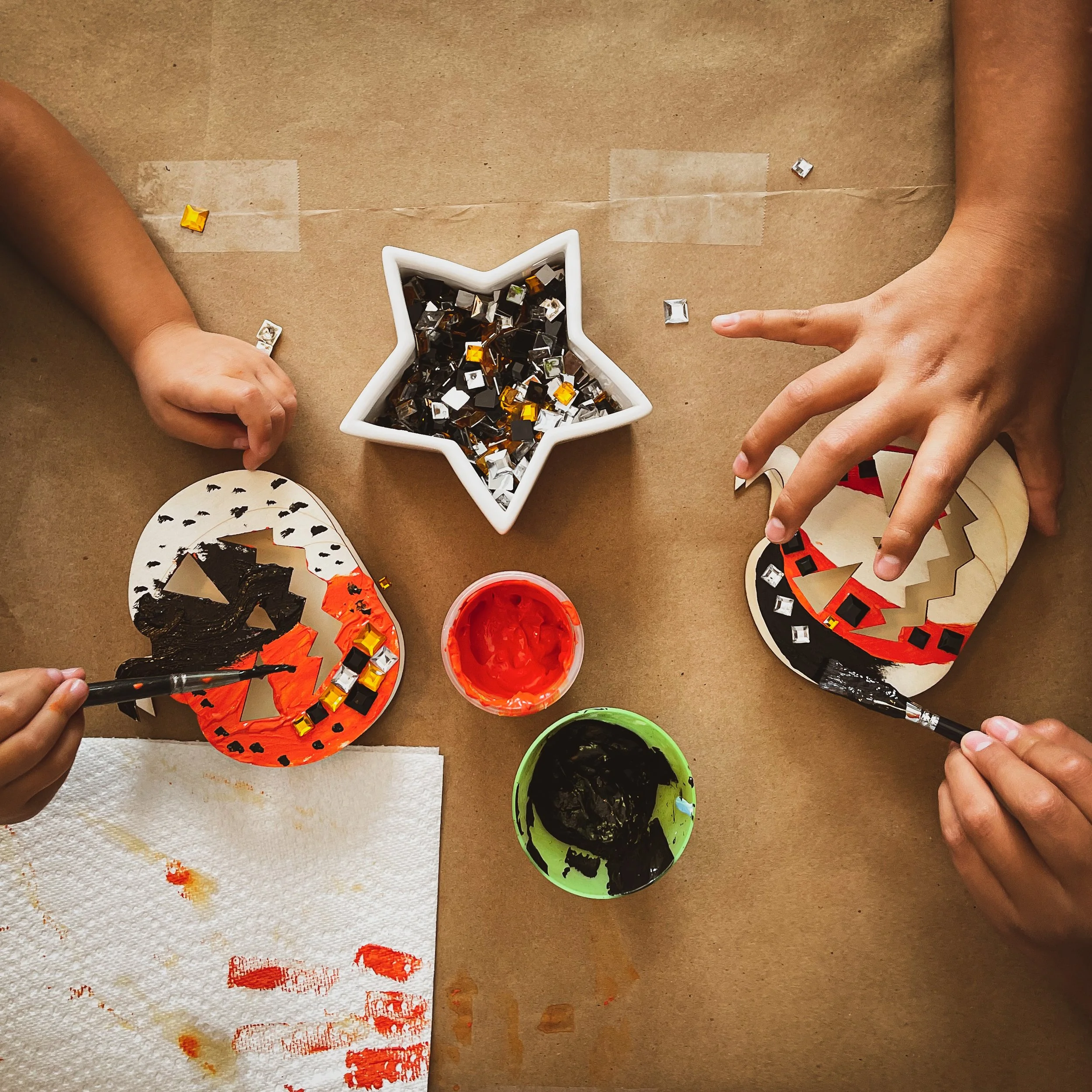 Children painting and decorating Halloween pumpkin masks with black, orange, and white paint, jewel embellishments, and craft supplies on a brown paper surface.