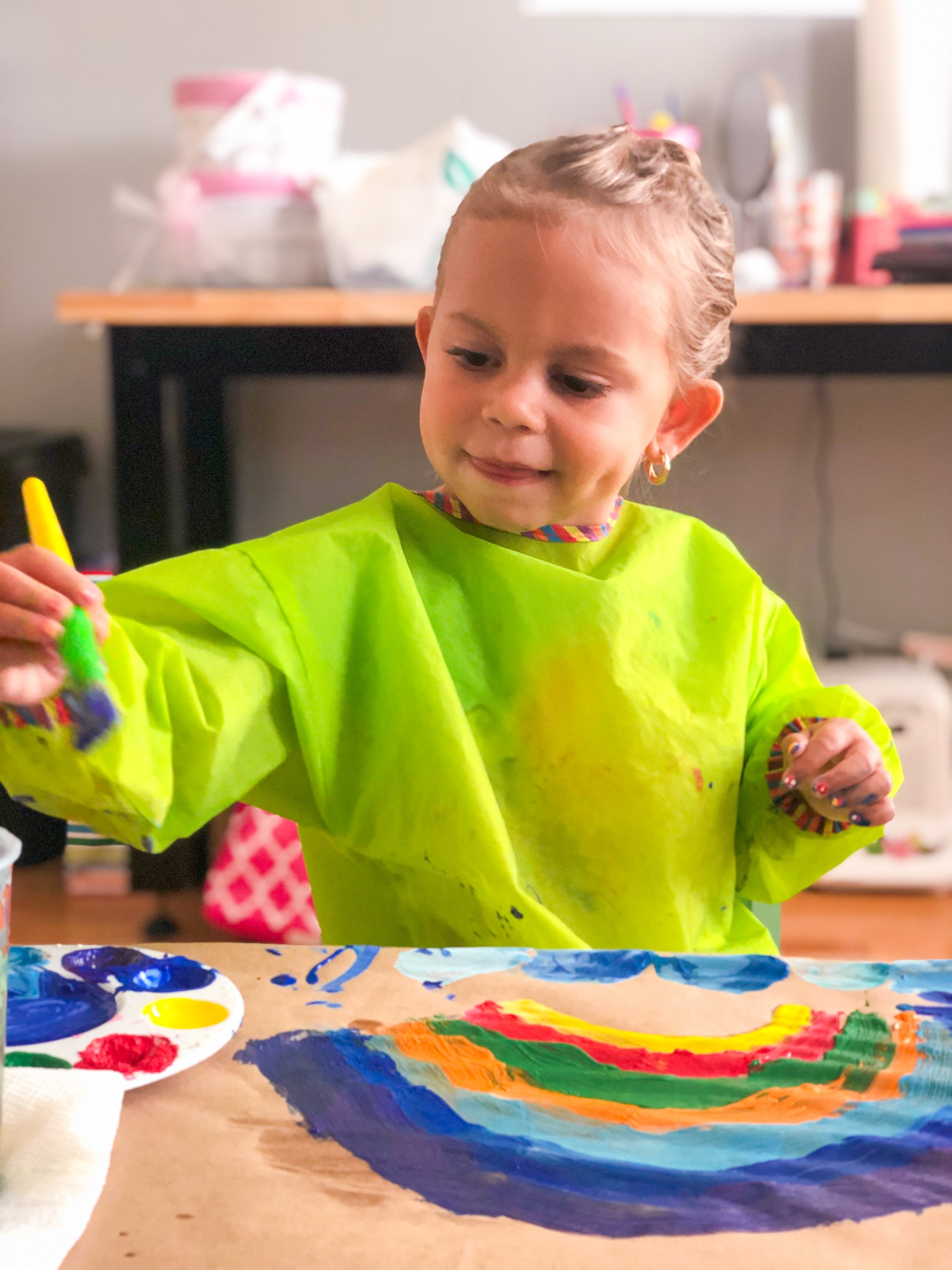 A young girl in a neon yellow smock paints a colorful rainbow on paper with finger paints, sitting at a table in a room that has art supplies on a shelf in the background.