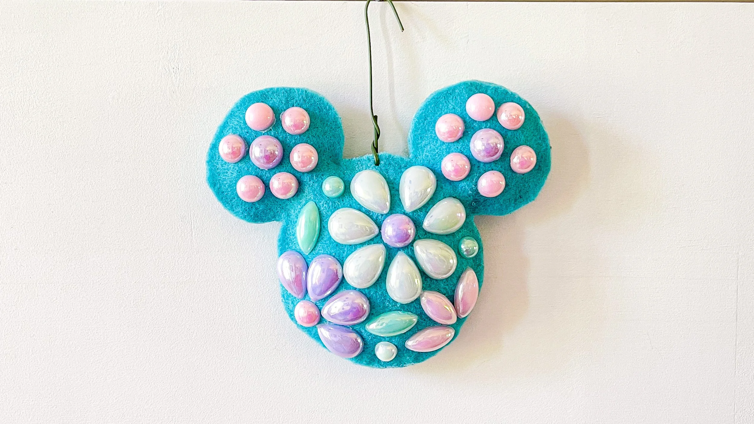 Decorative wall hanging in the shape of a Mickey Mouse head, made of turquoise felt with pink, white, purple, and iridescent beads and pearls attached.