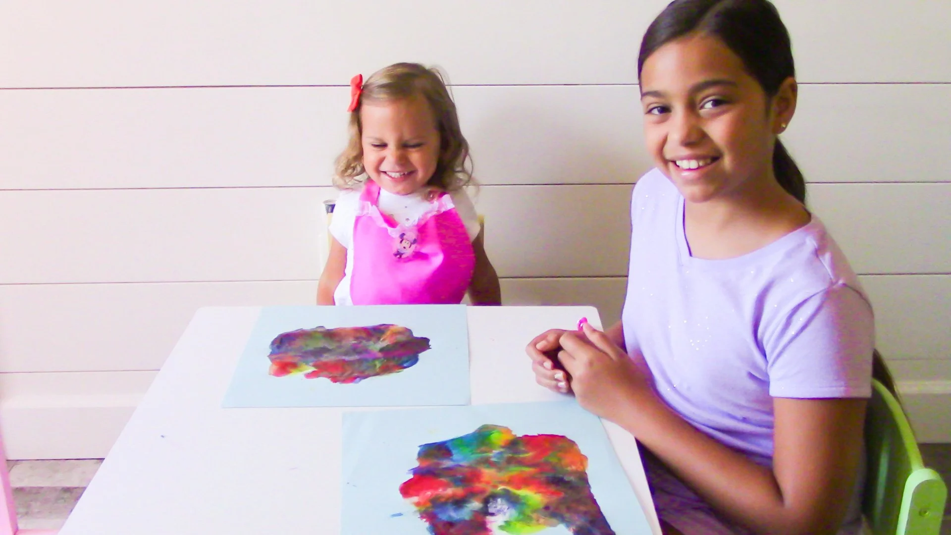 Two smiling young girls showing colorful art paintings on paper at a table.