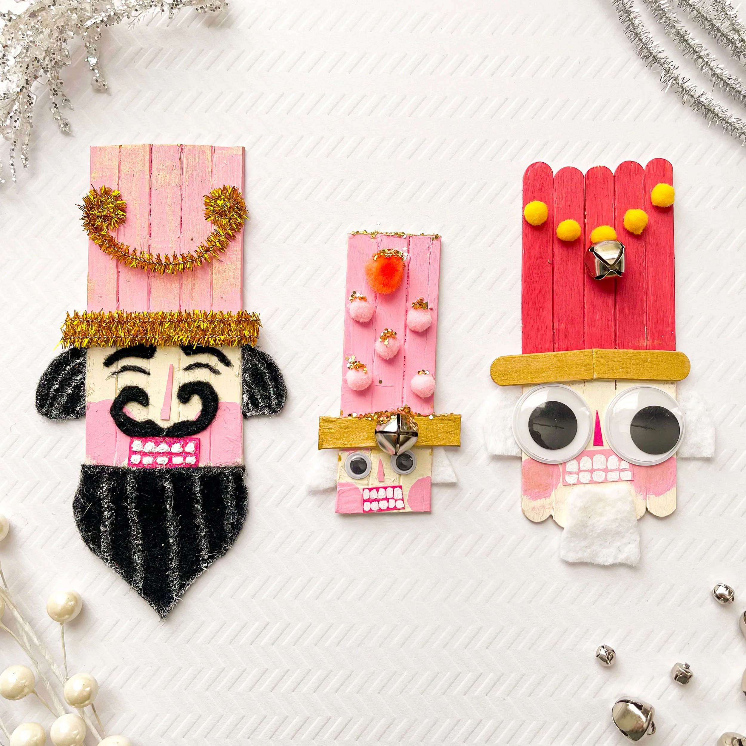 Three handmade holiday ornaments made from painted wood, pom-poms, googly eyes, bells, and other craft materials, designed to resemble whimsical faces with tall hats, on a white textured background with small silver jingle bells and white beads aroun