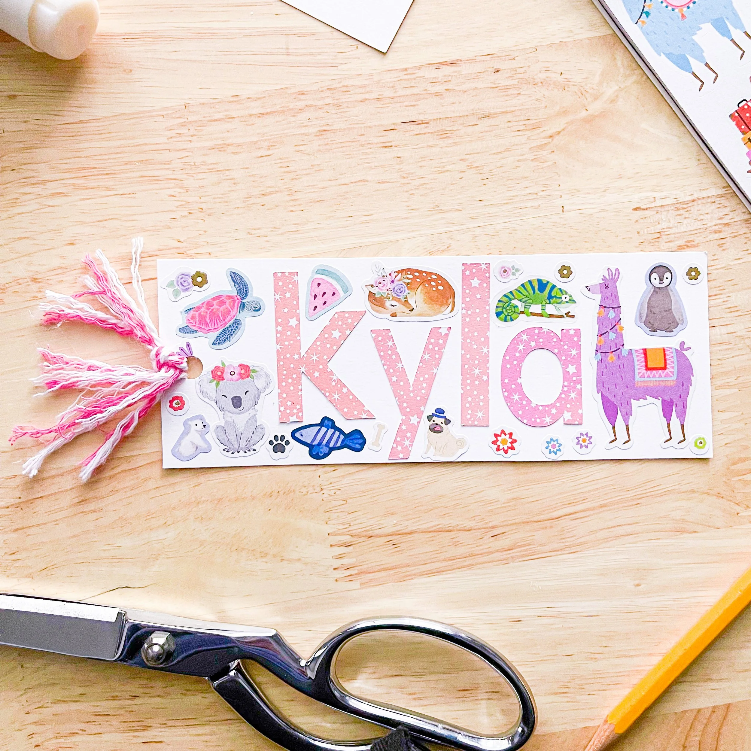 A handmade birthday banner with the name 'Kylia' in pink, decorated with animal stickers including a turtle, watermelon, lion, chameleon, penguin, llama, and fish, on a light wooden surface. There are pink and white tassels attached to one end of the