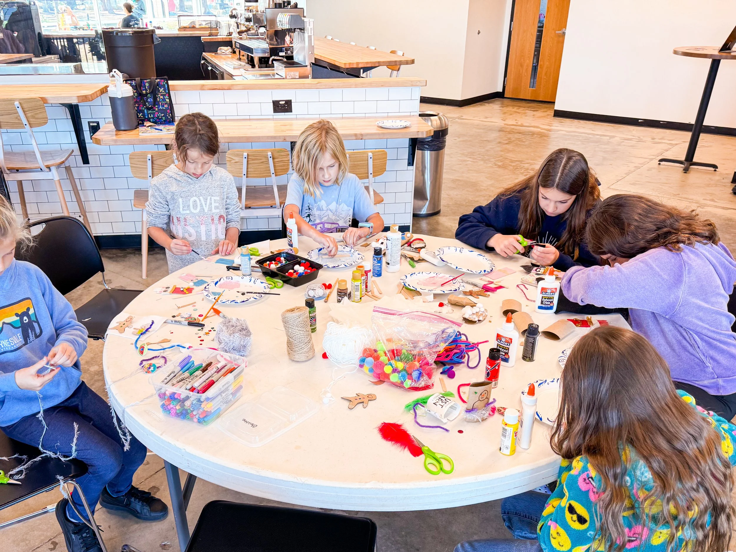 Children participating in arts and crafts activity at a round table with various craft supplies in a casual indoor setting.