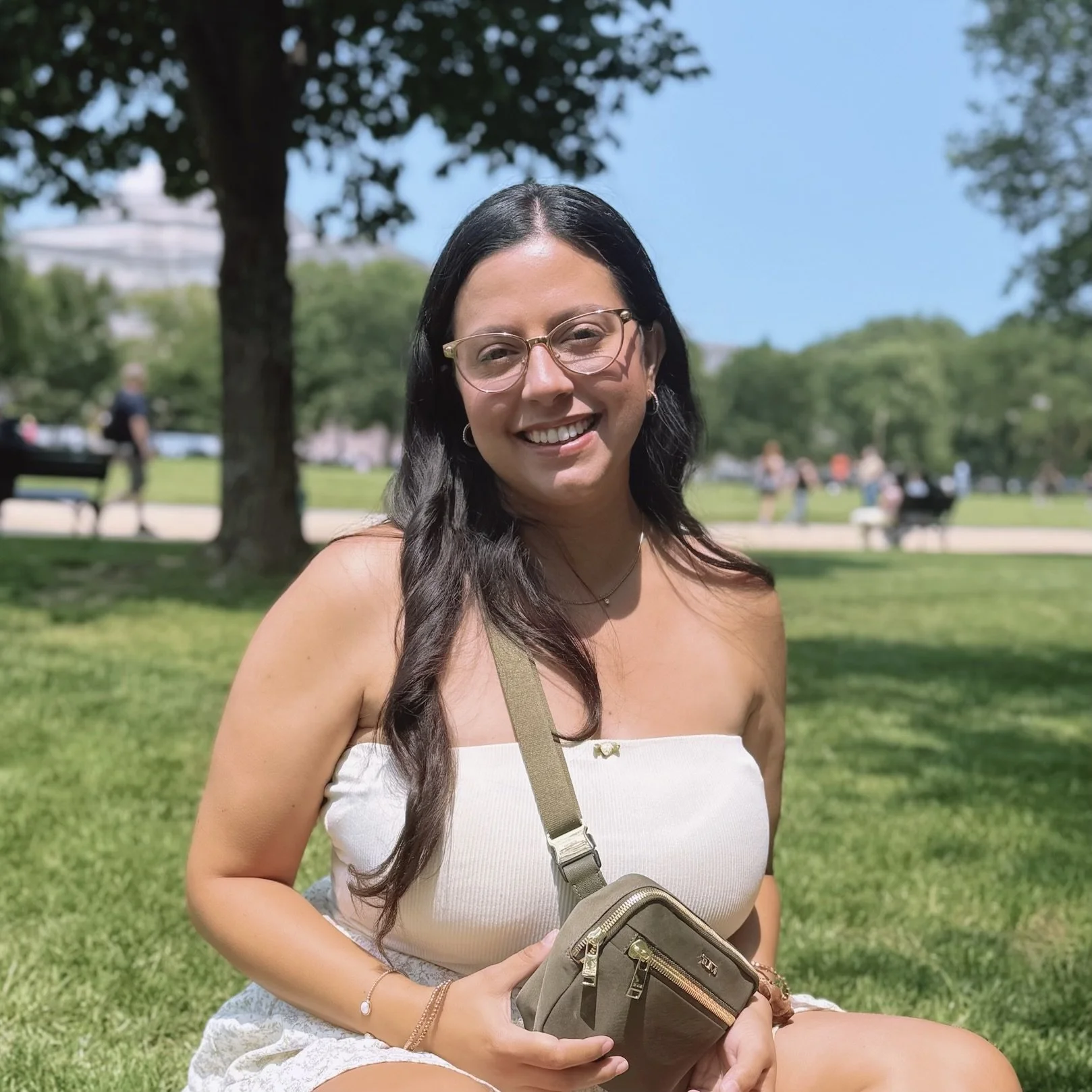 A woman sitting on the grass in a park, smiling, with trees and people in the background during a sunny day. Owner of Be Creative Tampa.