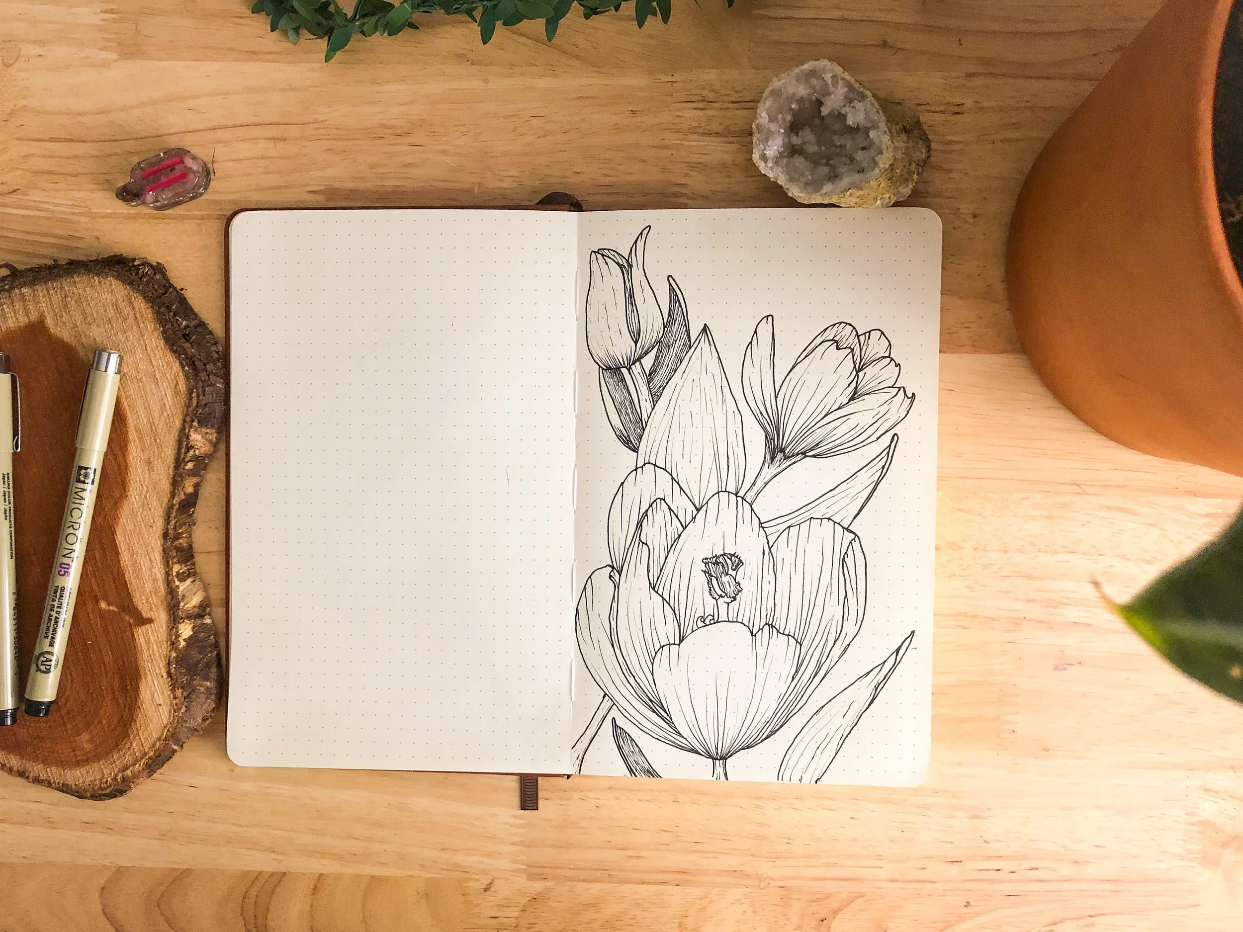 A sketch of tulips in a notebook surrounded by wooden, mineral, and ceramic objects on a wooden surface.