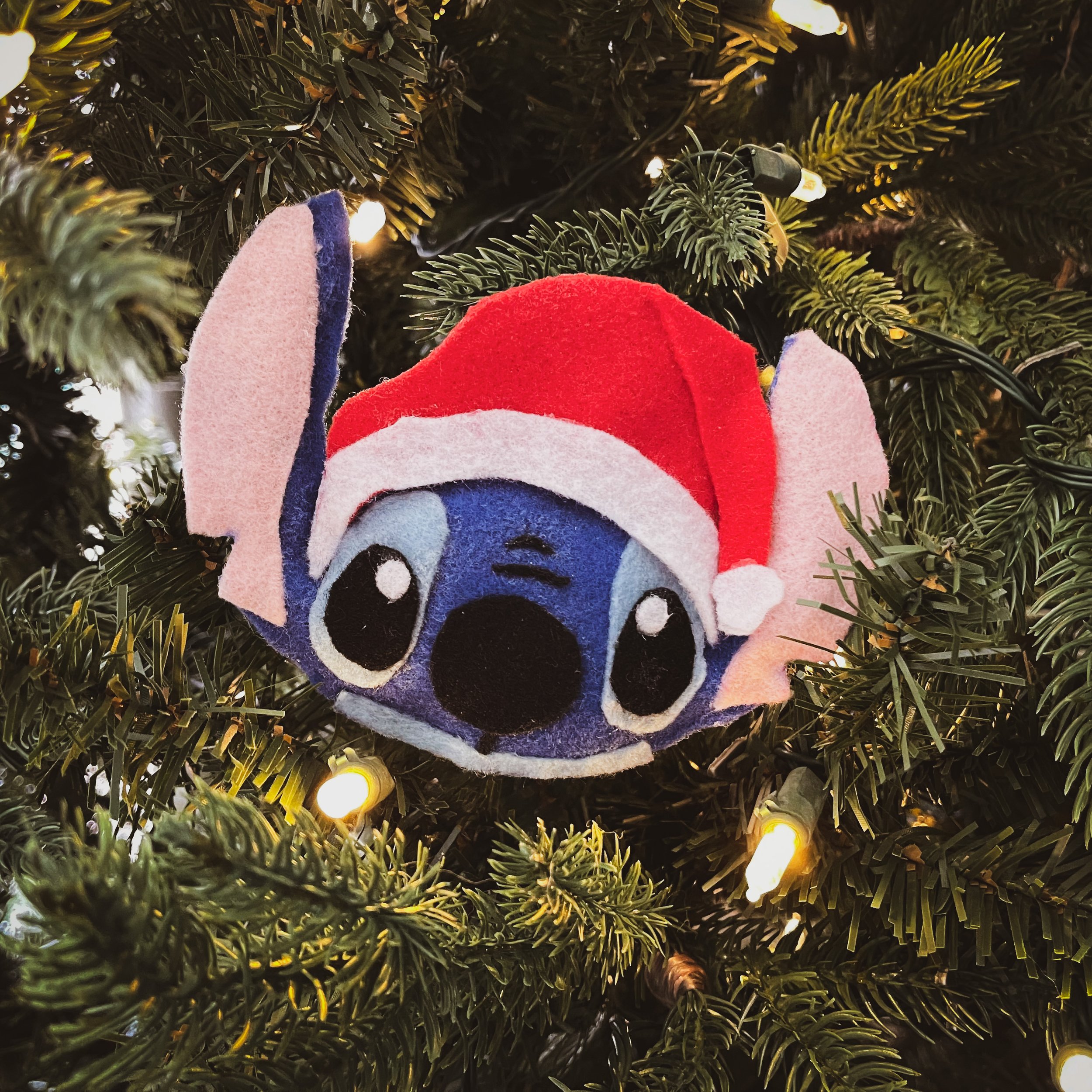 A plush ornament of Stitch from Lilo & Stitch wearing a Santa hat, hanging on a Christmas tree with lights.