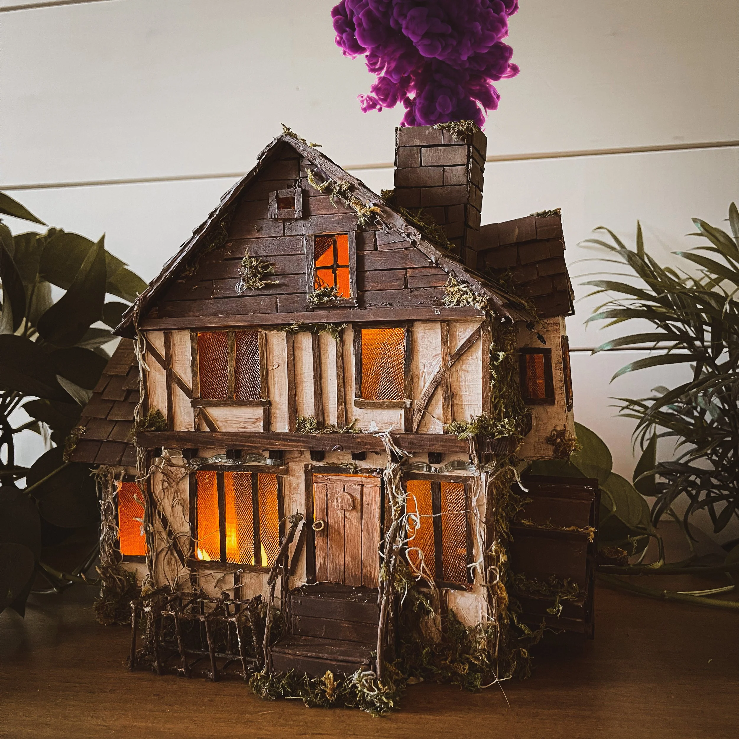 A miniature model of a two-story rustic house with a chimney emitting purple smoke, surrounded by green plants.