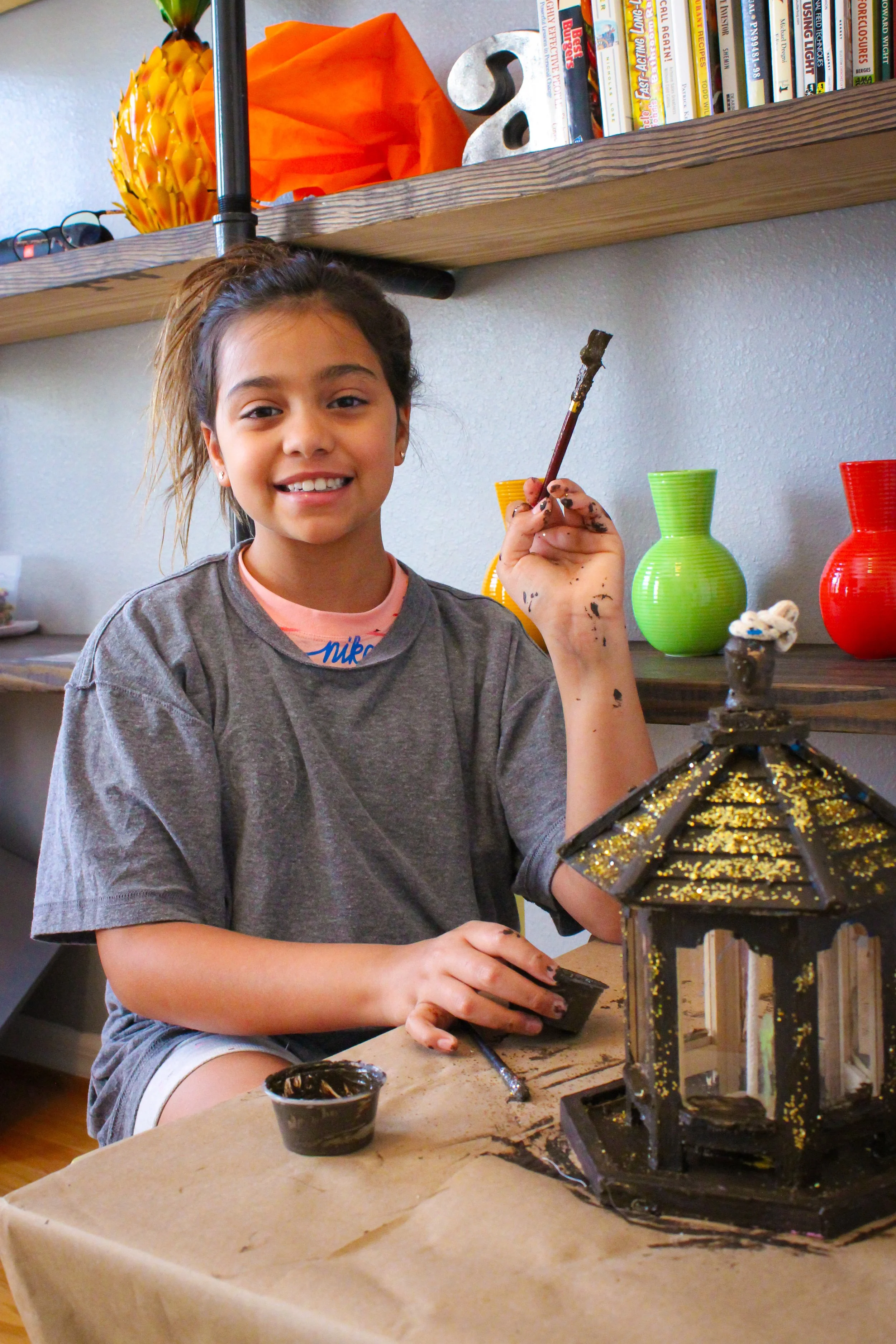 A young girl with messy hair and a gray t-shirt is painting a decorative lantern with black paint and gold accents. She is smiling and sitting at a table covered with brown paper, with painting supplies nearby. Books and colorful vases are on a shelf