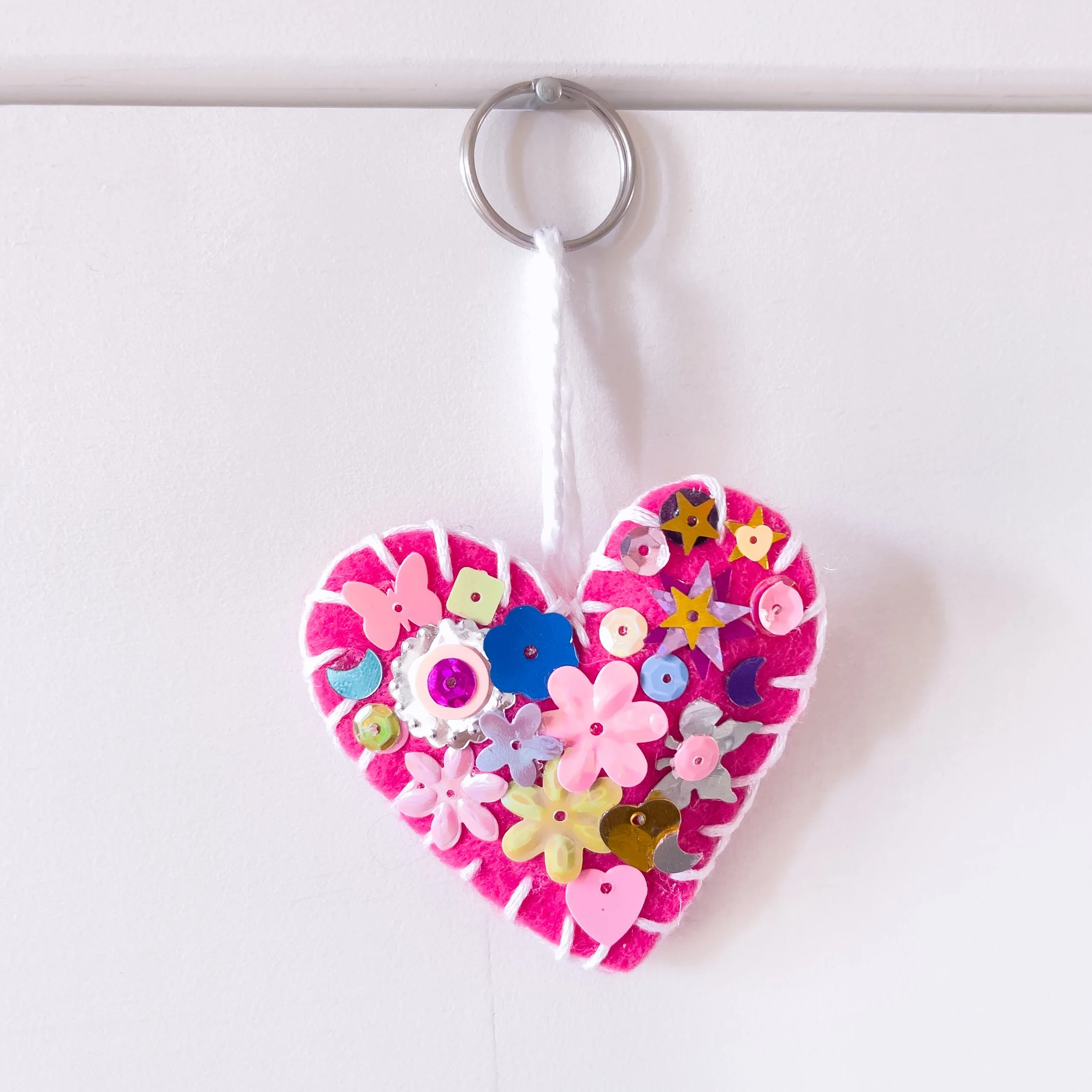 Pink felt heart-shaped hanging decoration decorated with colorful sequins, beads, and small appliqué shapes, hanging from a clear plastic loop attached to a white string on a white wall.