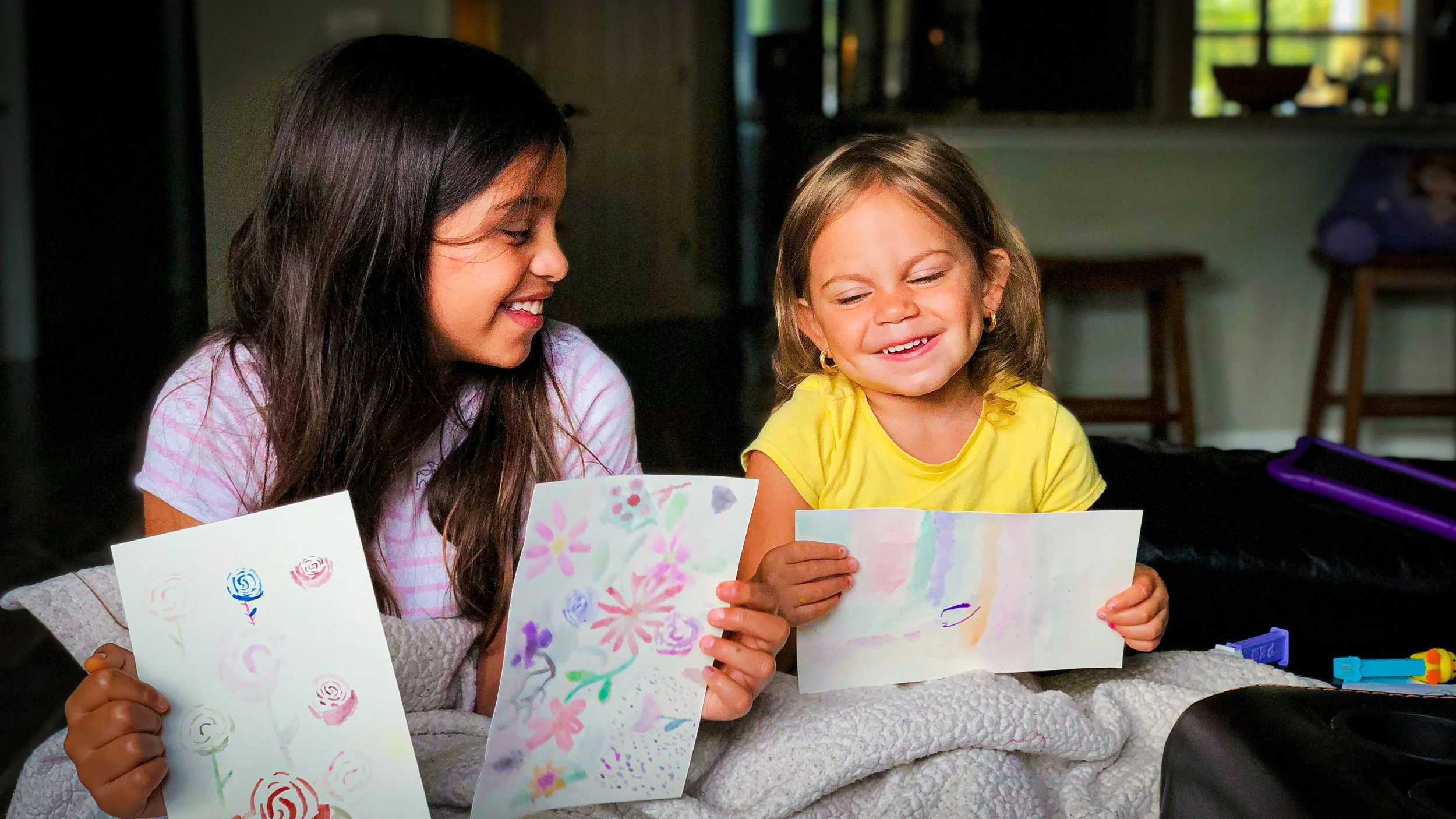 Two young girls sitting on a bed, smiling and holding colorful drawings they made, with a cozy room in the background.