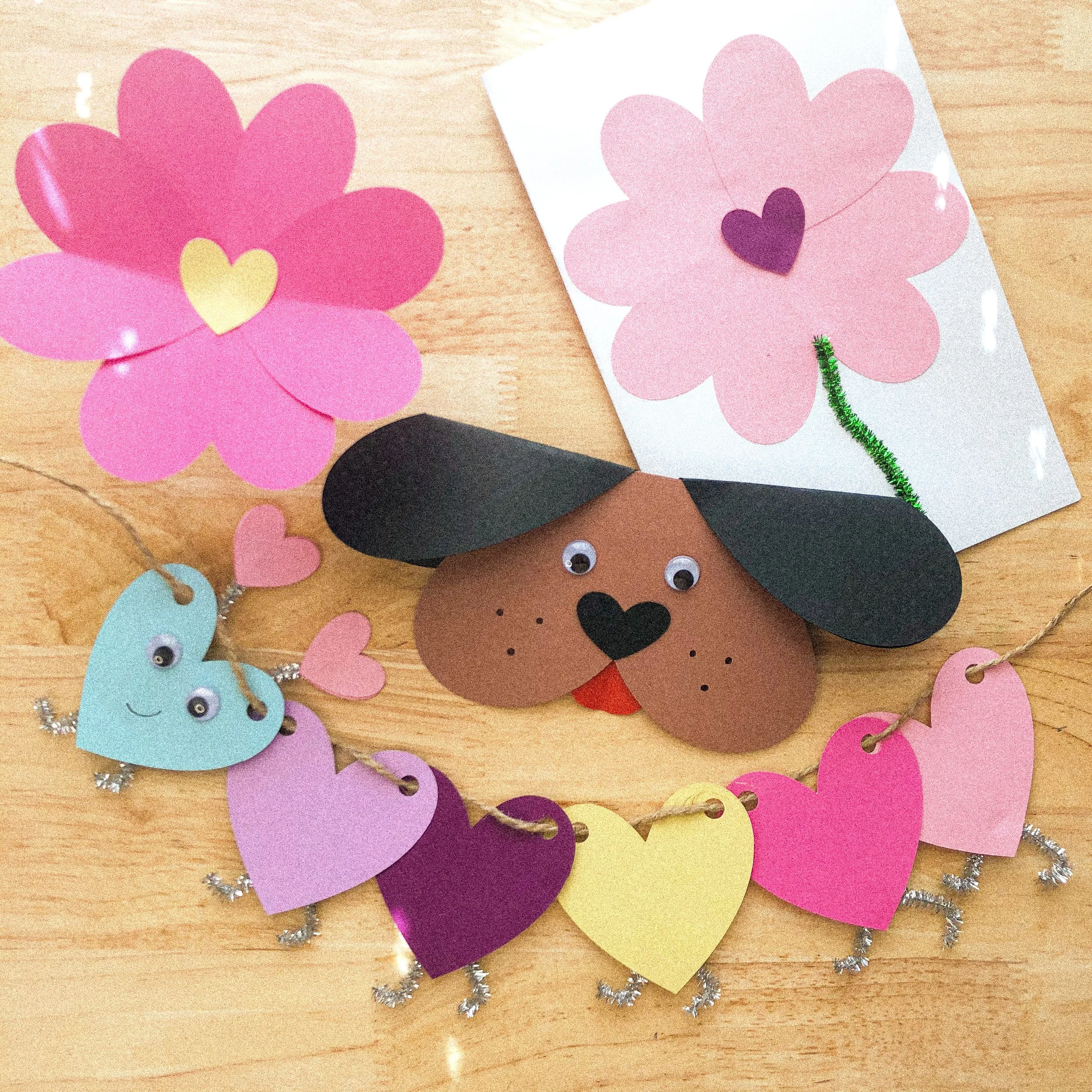 Decorative craft items including paper flower, heart-shaped and animal shapes with googly eyes, and a garland of paper hearts on a wooden surface
