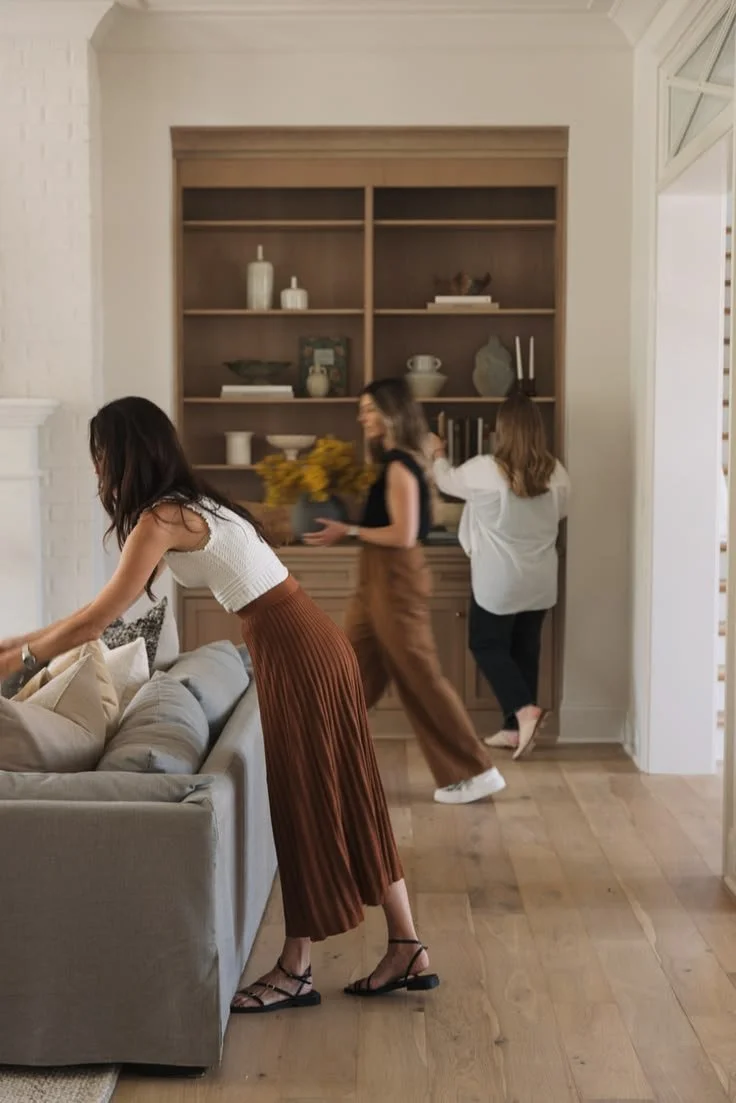 Three women are in a living room, with one woman arranging pillows on a beige sofa, another woman carrying a bouquet of yellow flowers, and the third woman standing by a wooden bookshelf with decorative items.