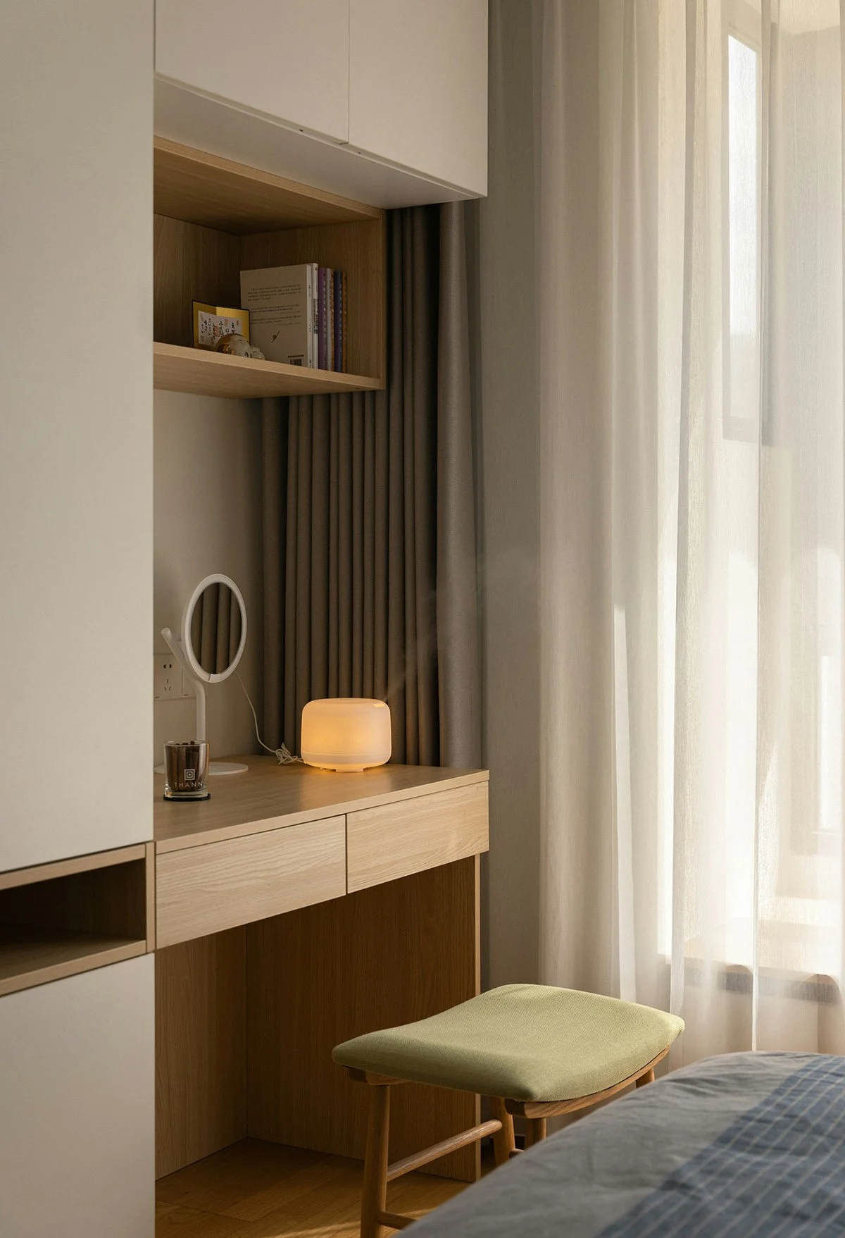 A bedroom corner with a wooden desk, a small lamp, a round mirror, books on a shelf, and a green cushioned stool. Light filters through sheer curtains.