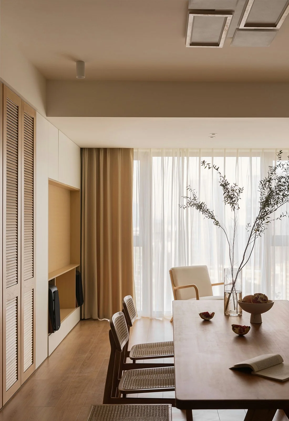 A modern dining room with wooden furniture, a large window with sheer curtains, a beige armchair, a glass vase with branches on the table, and bowls of fruit.