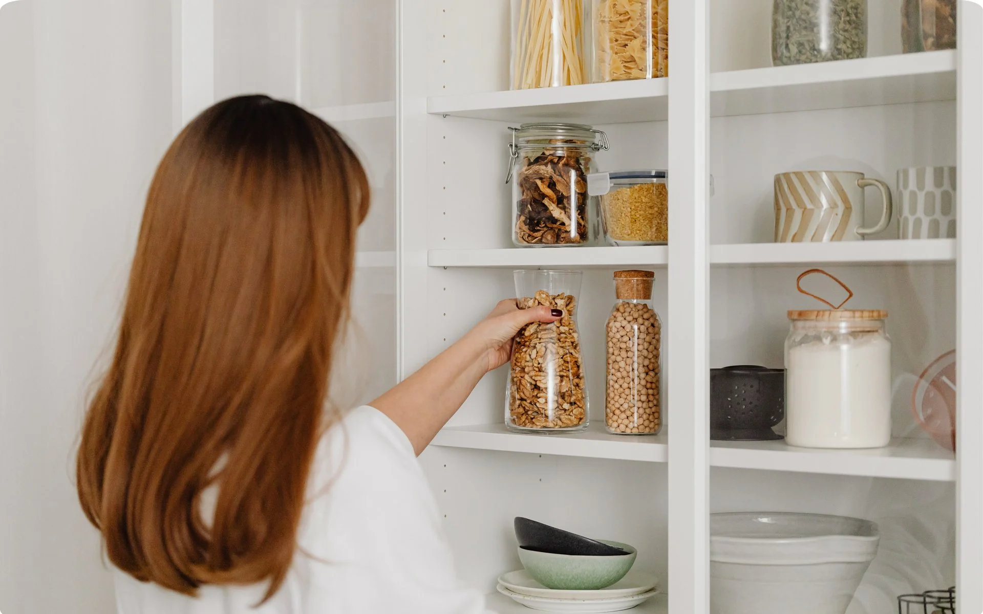 Woman with long red hair reaching into a glass jar of mixed nuts in a white kitchen pantry.