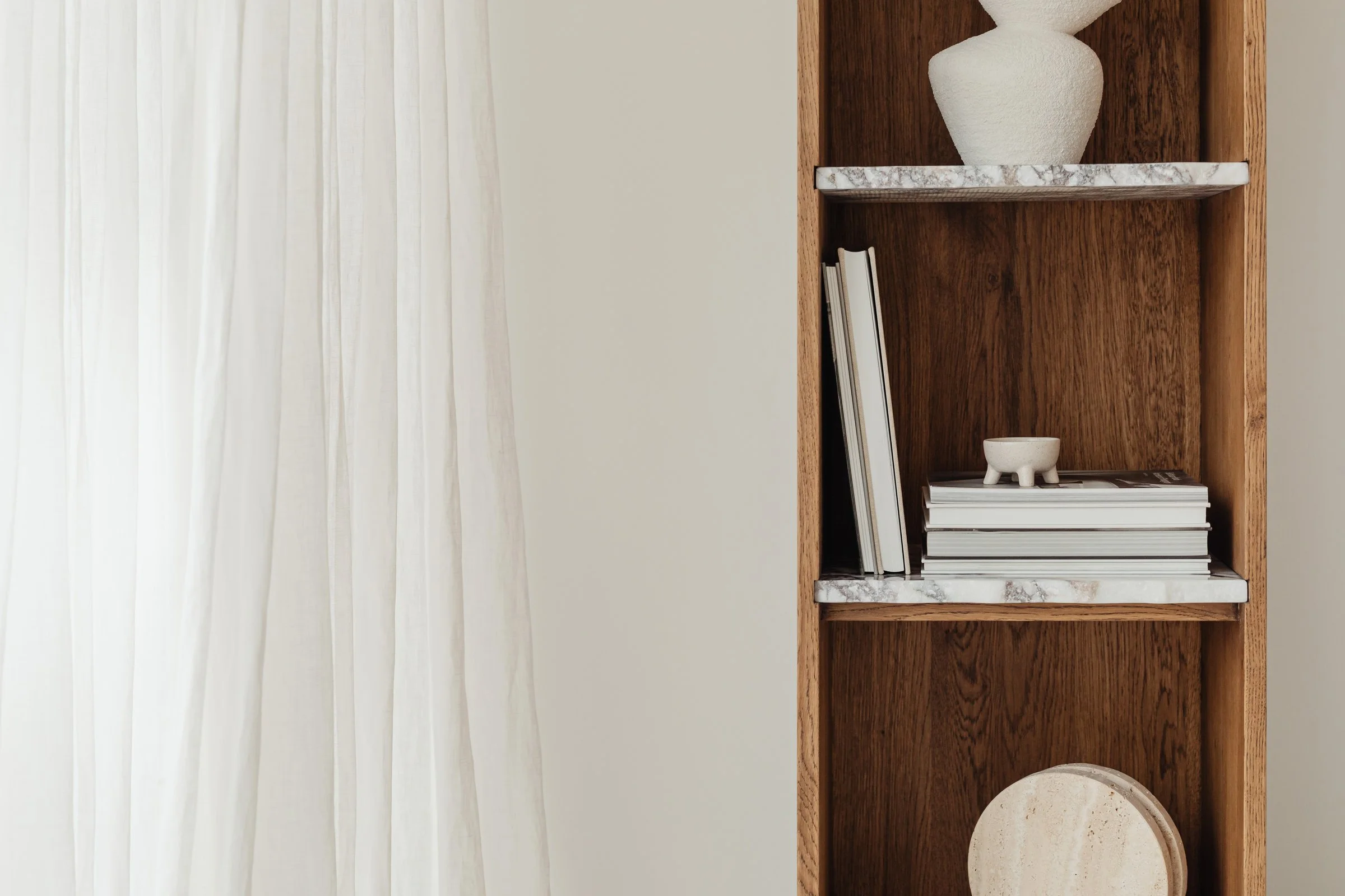 A wooden bookshelf with decorative objects and books, next to light-colored curtains.