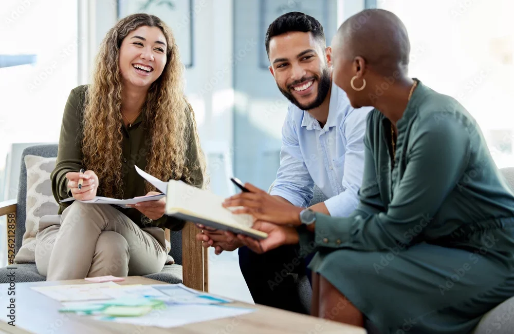 Three diverse young adults smiling and talking together in a bright, modern office space, with notebooks and papers on a coffee table.