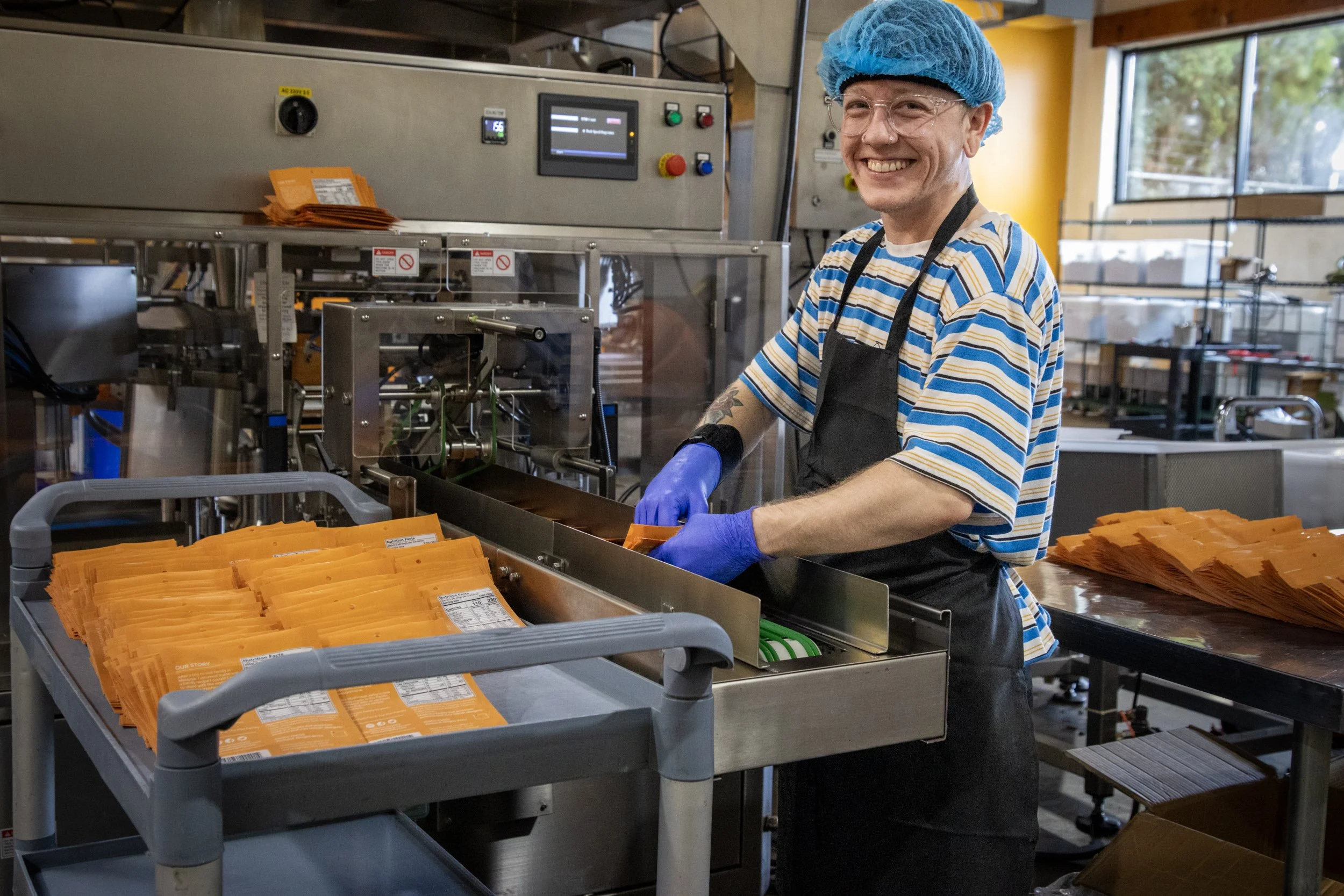 A smiling man in a striped T-shirt, blue hairnet, glasses, and gloves working in a food packaging facility. He is standing at a conveyor belt with orange food packaging pouches, with machinery and a window in the background.