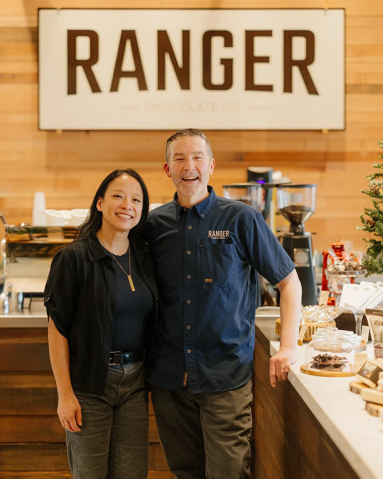 Two people, a woman and a man, standing inside a coffee shop with a large 'RANGER' sign behind them, smiling at the camera.