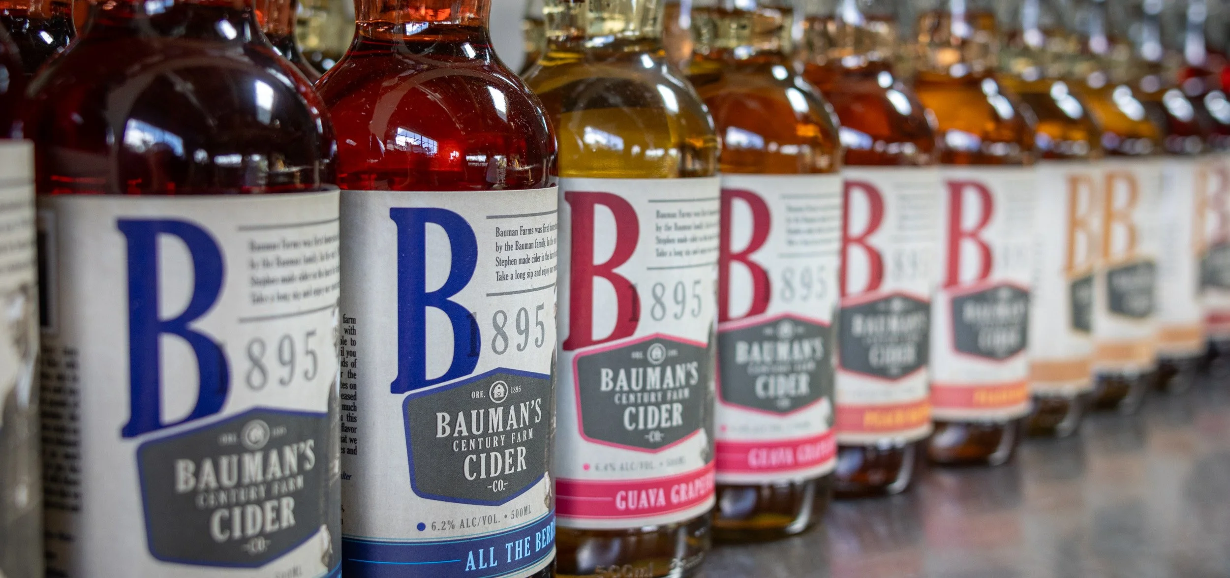 Bottles of Bauman's Century Farm cider on a store shelf, with labels indicating different flavors such as Guava Grape.