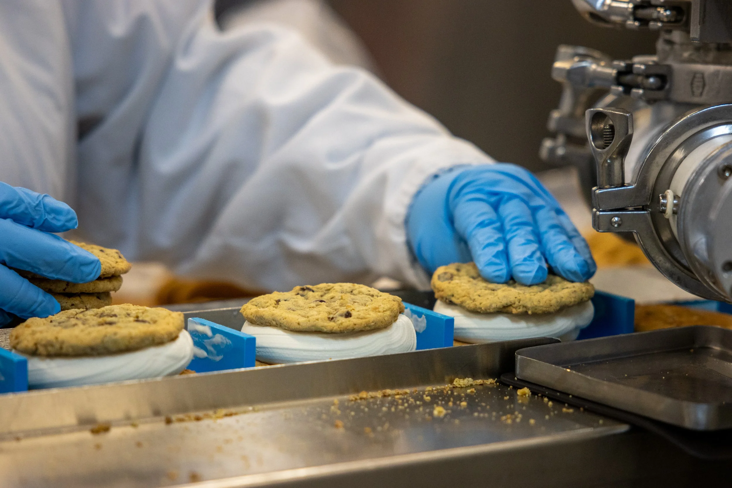 A person wearing white protective clothing and blue gloves is placing freshly baked chocolate chip cookies onto a cooling rack in a bakery or food production facility.