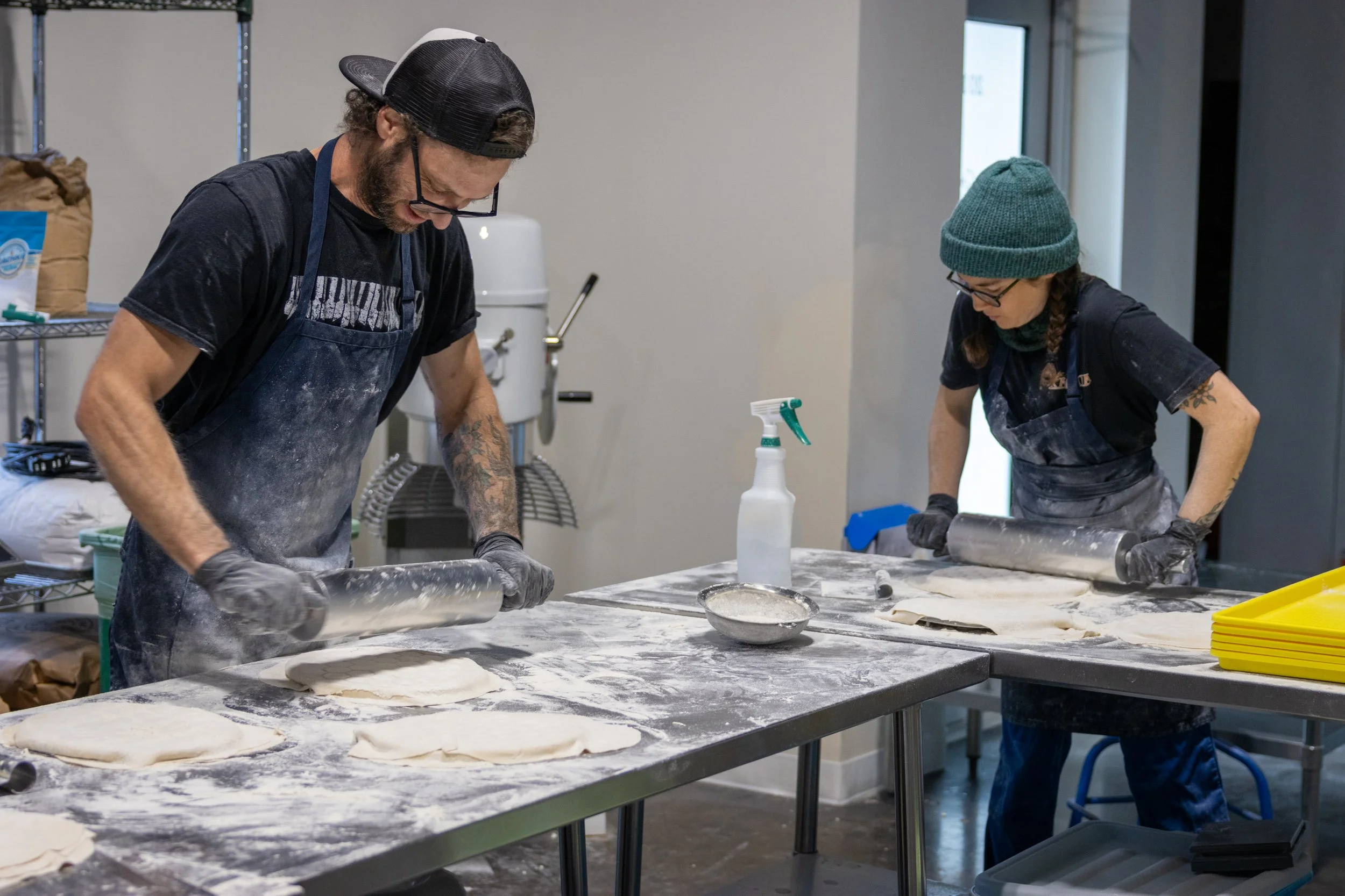 Two people are working in a bakery, rolling out dough on a floured table. One person, with curly hair and glasses, is wearing a black cap backwards and a black apron. The other person, with long hair in braids, is wearing a green beanie and glasses. There are various baking supplies on the table, including a spray bottle and a bowl of flour.