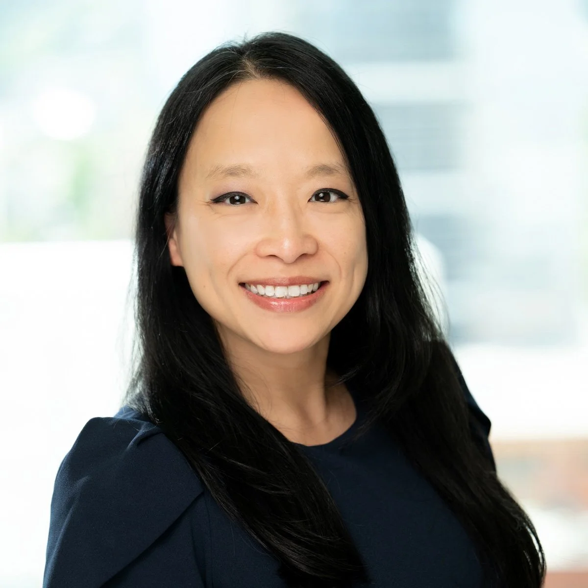 A woman with long black hair wearing a navy blue top, smiling in front of a blurred bright background.