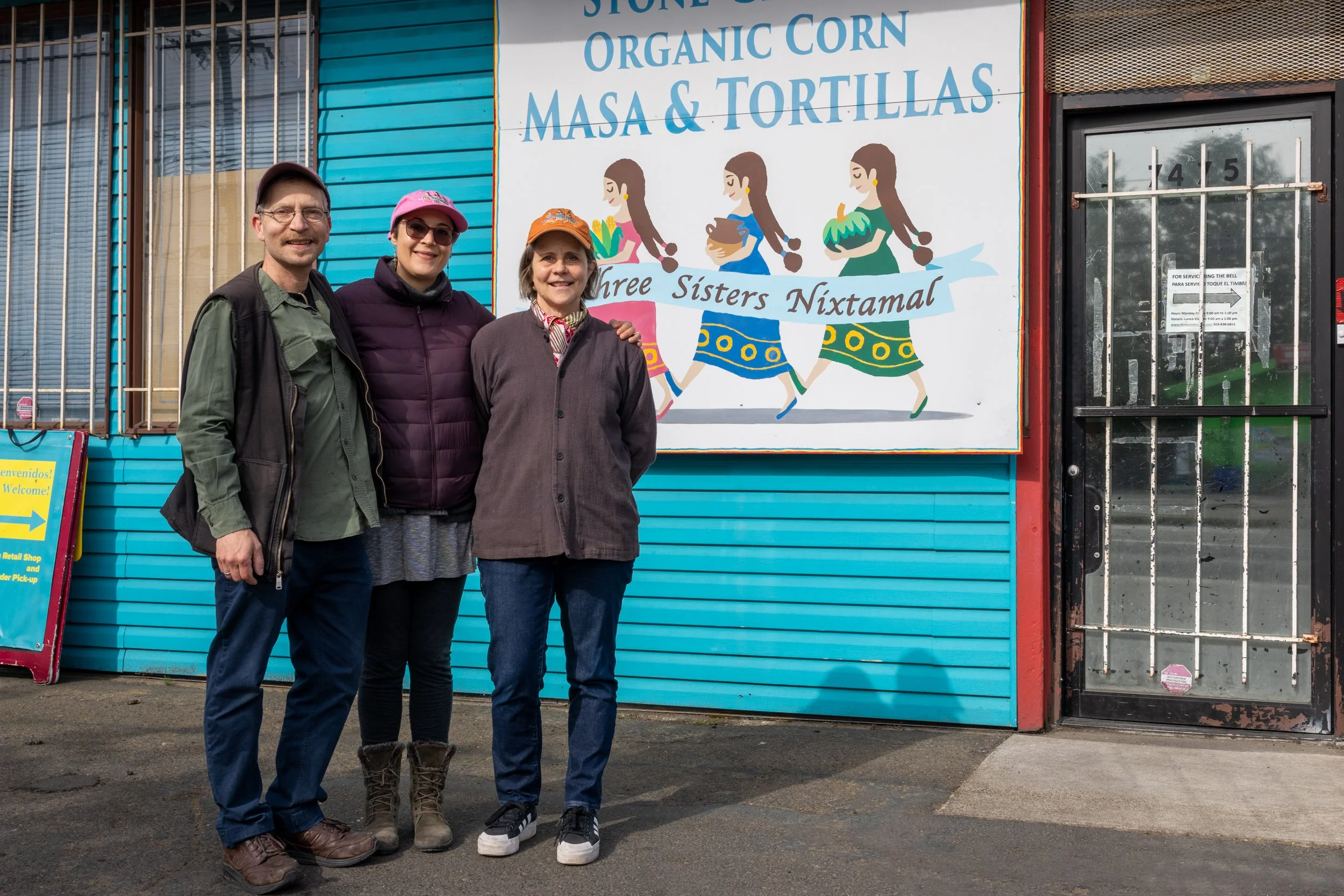 Three people standing outside a blue building with a large sign that reads 'Stone Organic Corn Masa & Tortillas' and features illustrations of women holding corn and tortillas.
