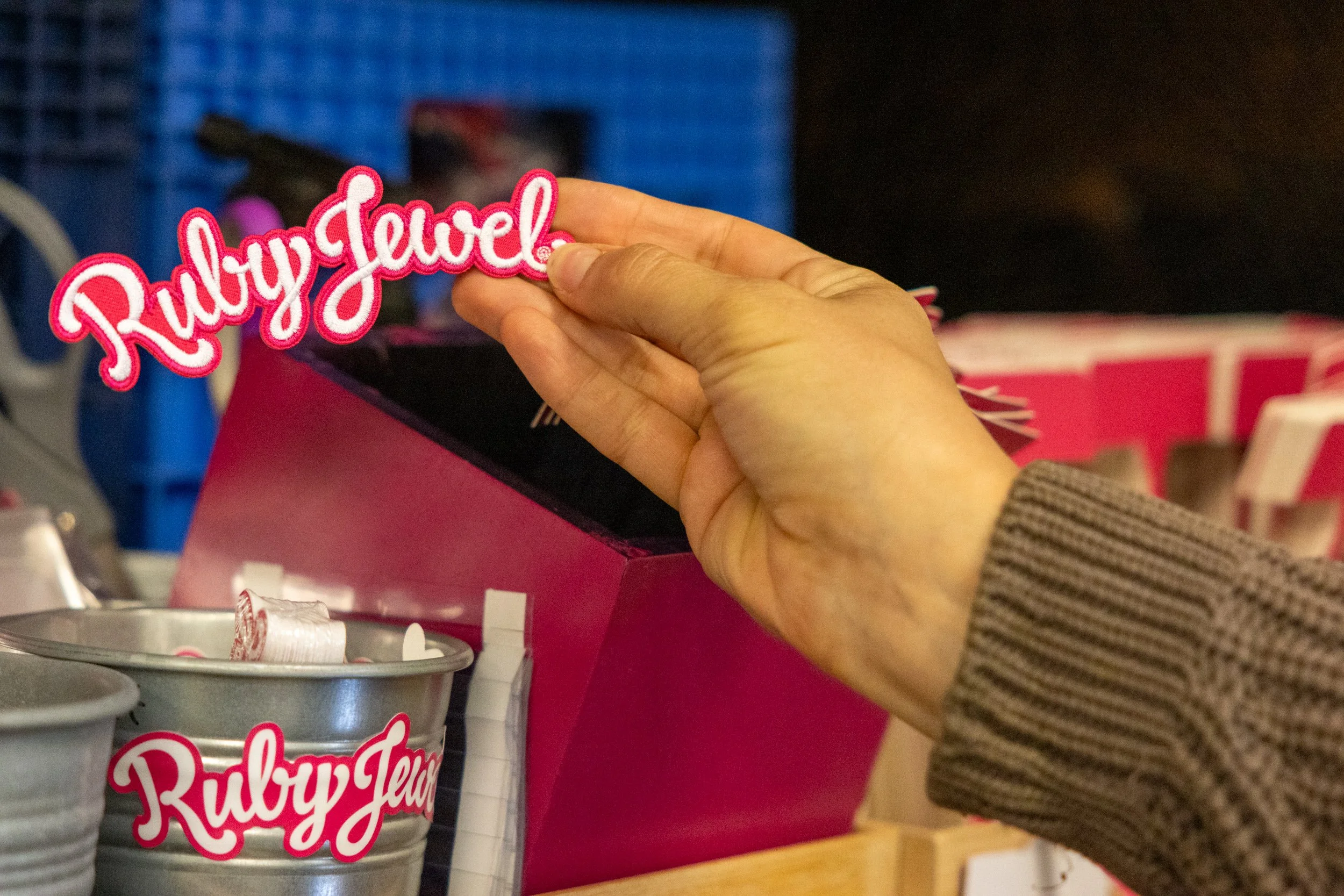 A hand holding a pink and white embroidered patch that says 'Rubygirl!' in front of a red box with cans of Kulpy Jelly and other canned goods on a store shelf.