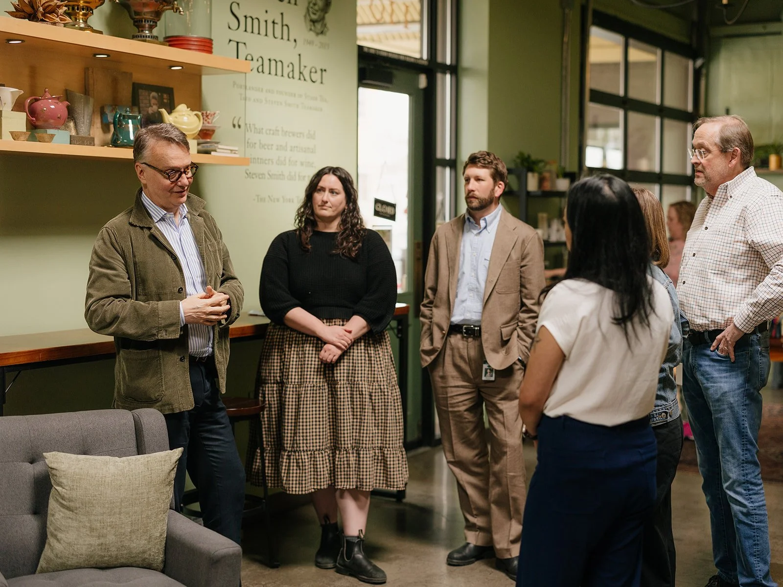 A group of six people having a discussion or debate in a room with green walls and shelves with teapots and books, near a sign that says "Smith, Teamaker."