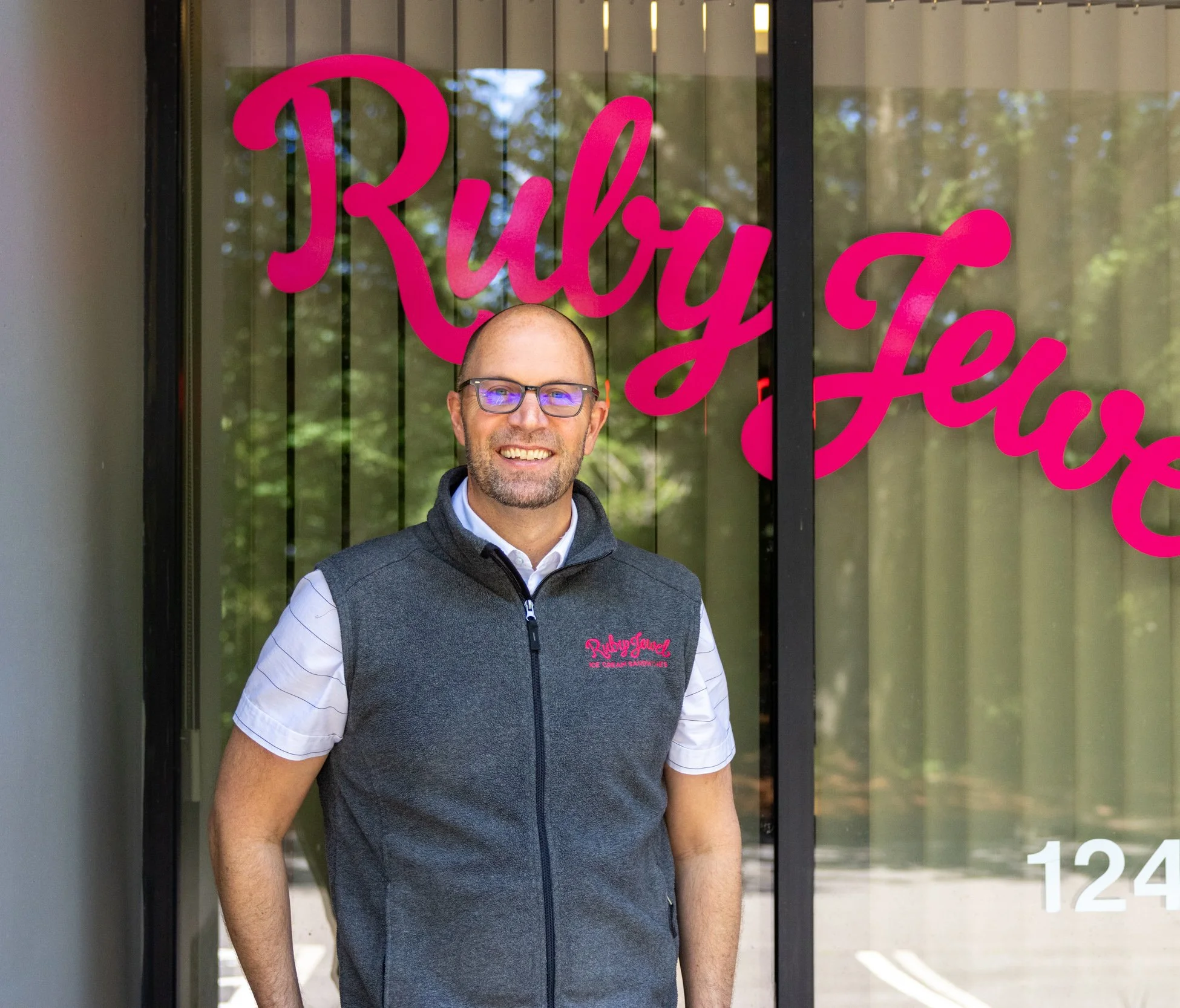 A smiling man with glasses wearing a gray vest over a white shirt, standing outside near a window with pink text that says 'Ruby'.