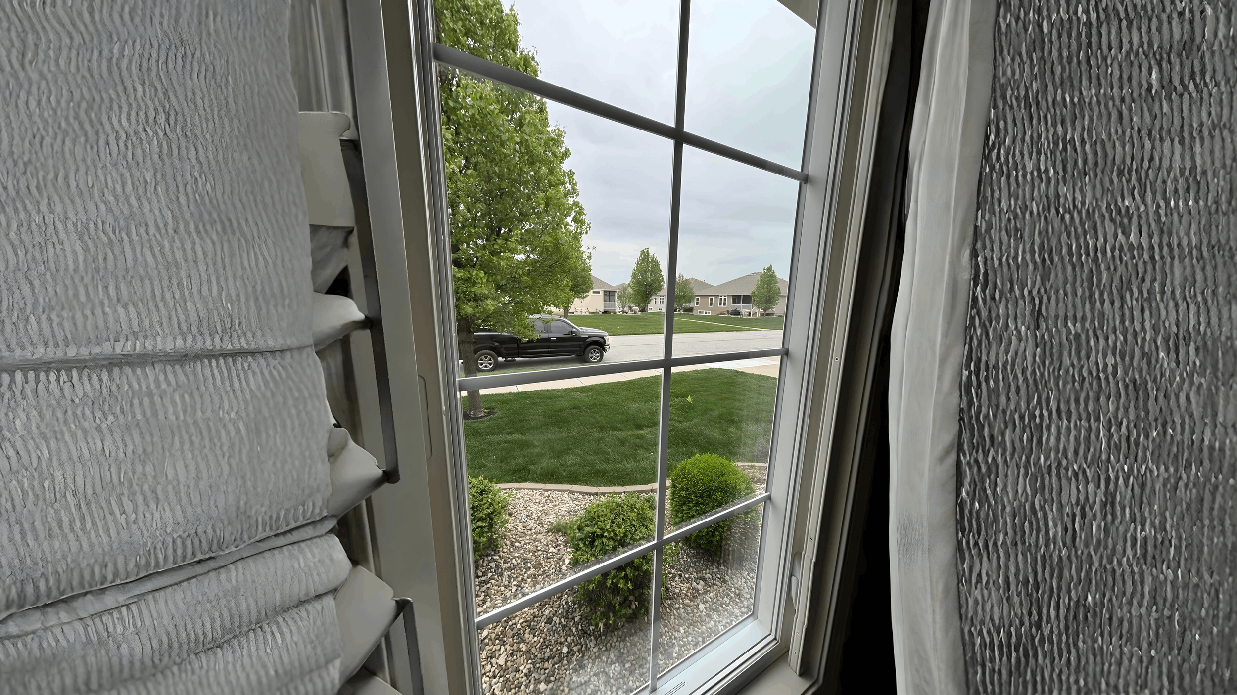 View through a window with white blinds partially pulled up, showing a suburban street with a black pickup truck, green lawn with small bushes, and houses in the background with trees and an overcast sky.