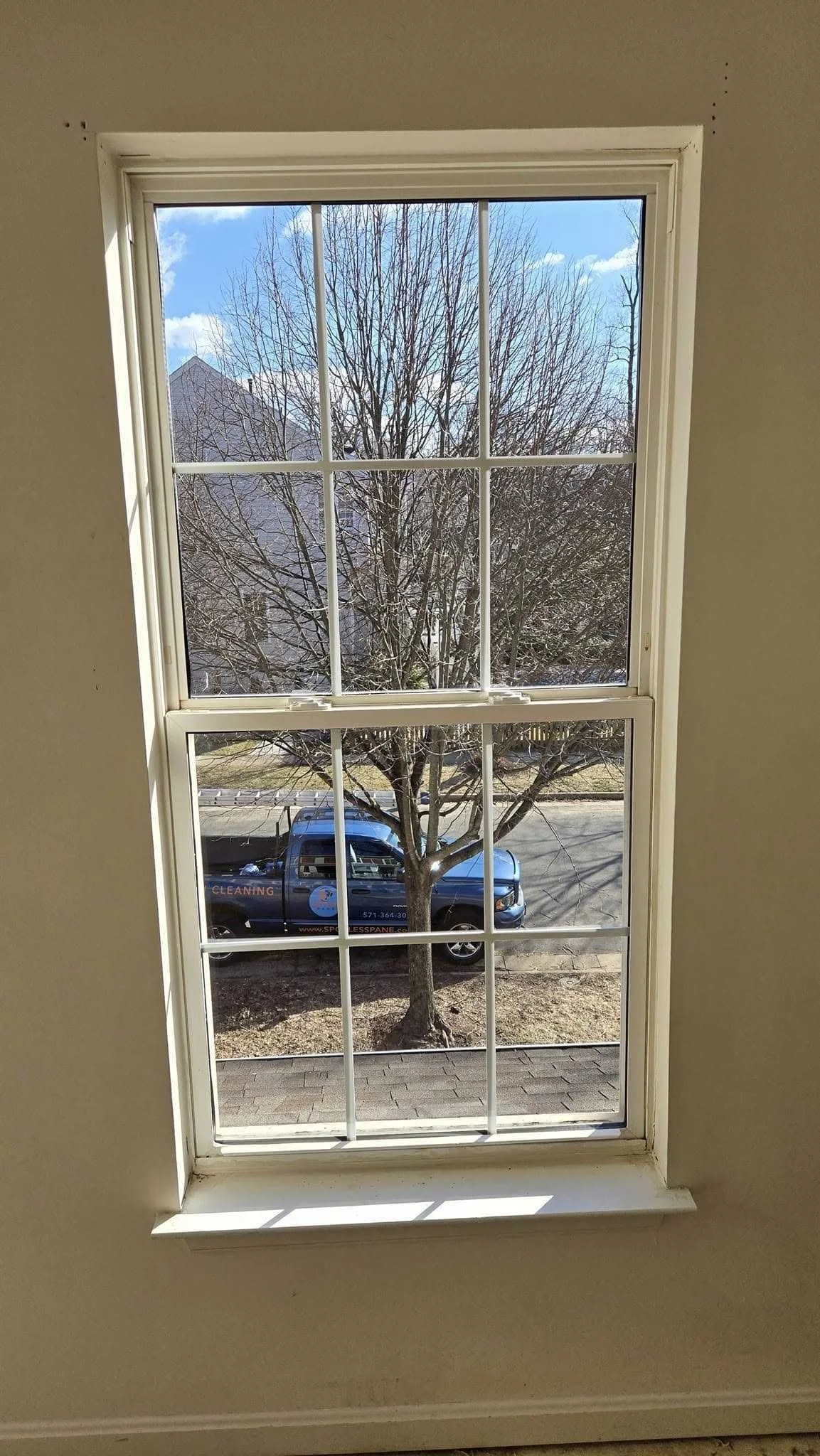 View from inside a room through a double-hung window showing a tree with bare branches, a parked blue truck on the street, and a clear blue sky with some clouds.