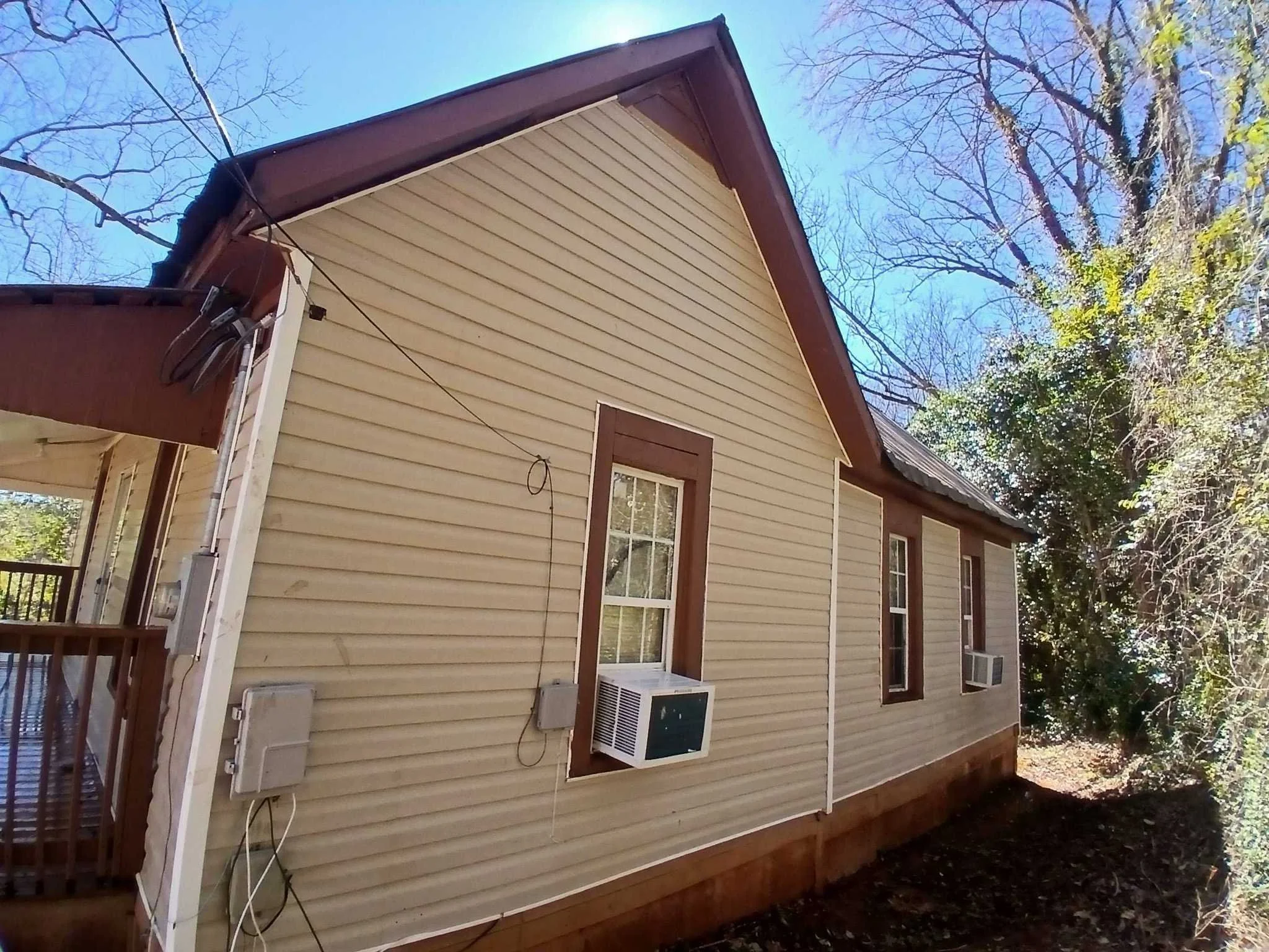 Side view of a beige, vinyl-sided house with three windows with brown trim, two of which have window air conditioning units. The house has a brown roof, and there's a porch to the left. Surrounding trees have no leaves, suggesting fall or winter.