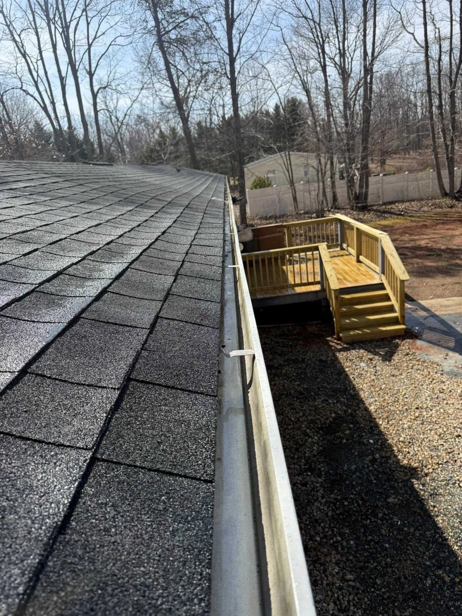 Close-up of a house roof with black shingles and a gutter, with a backyard patio and a wooden deck with stairs, surrounded by trees and a fence in the background.