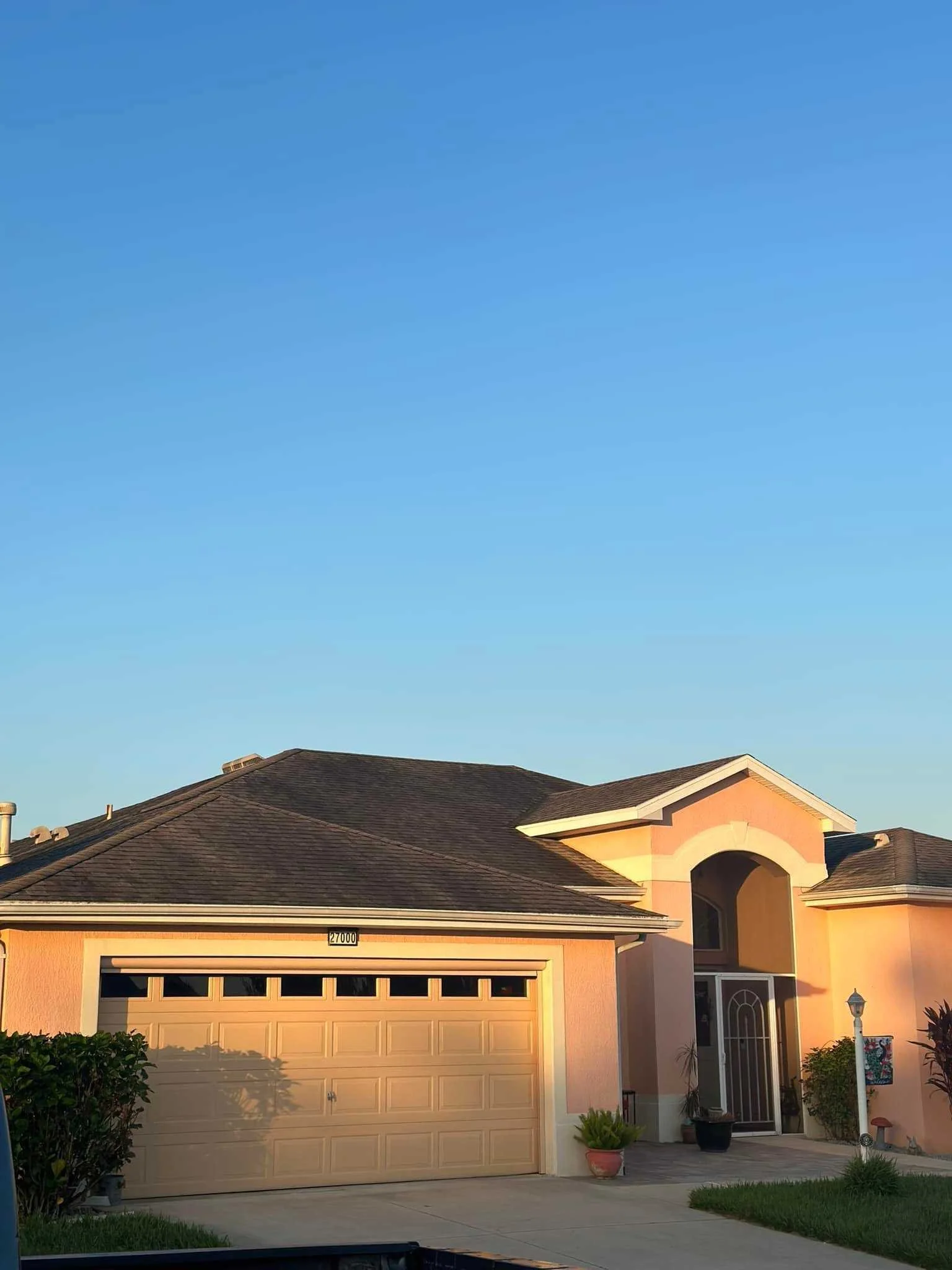 Front view of a peach-colored house with a brown shingle roof, a two-car garage, and a small front porch. The house has potted plants and a lamp post near the entrance, with a clear blue sky in the background.