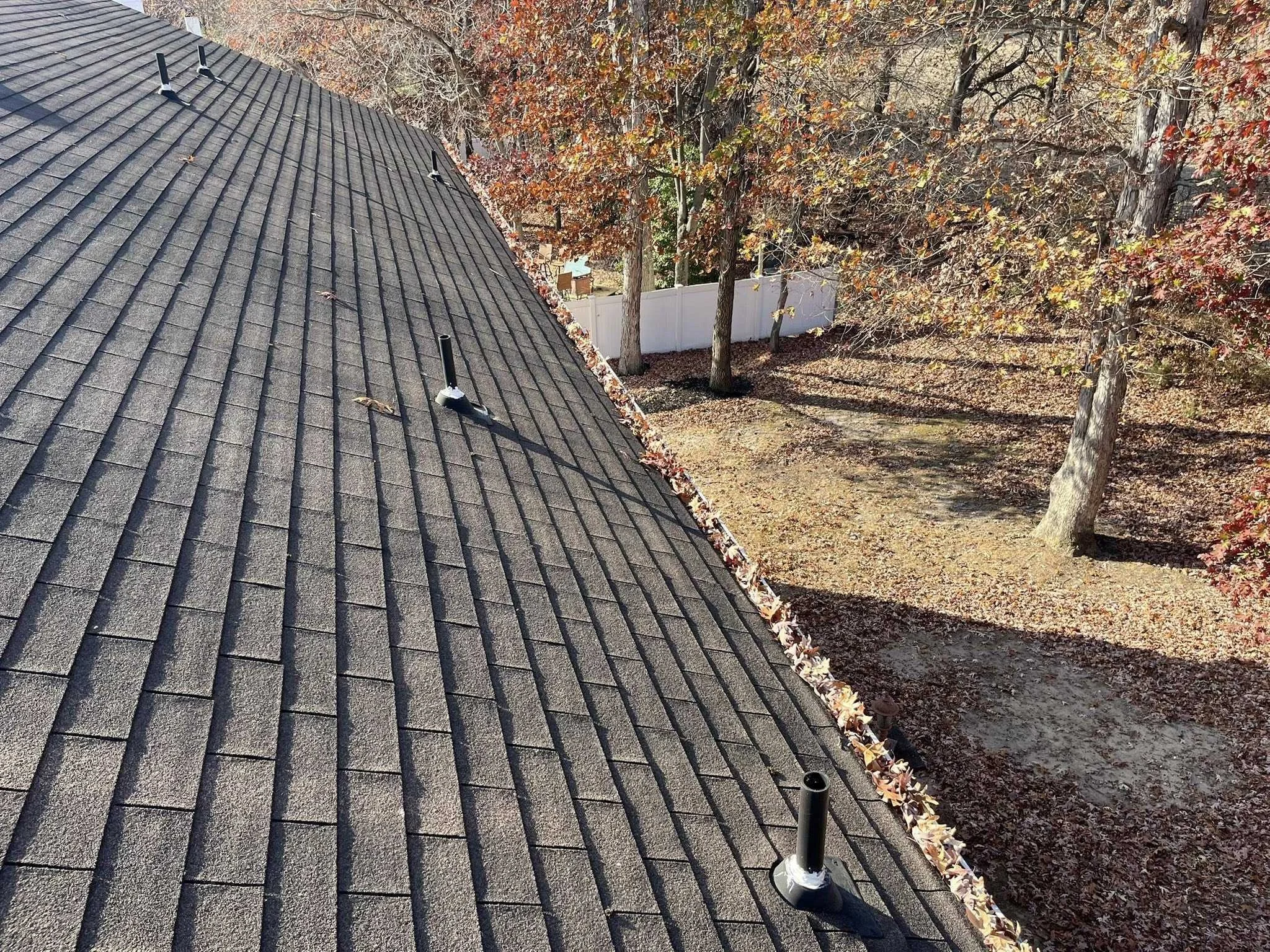 View of a shingled rooftop with four vents, overlooking a backyard with trees, fallen leaves, and a white fence.