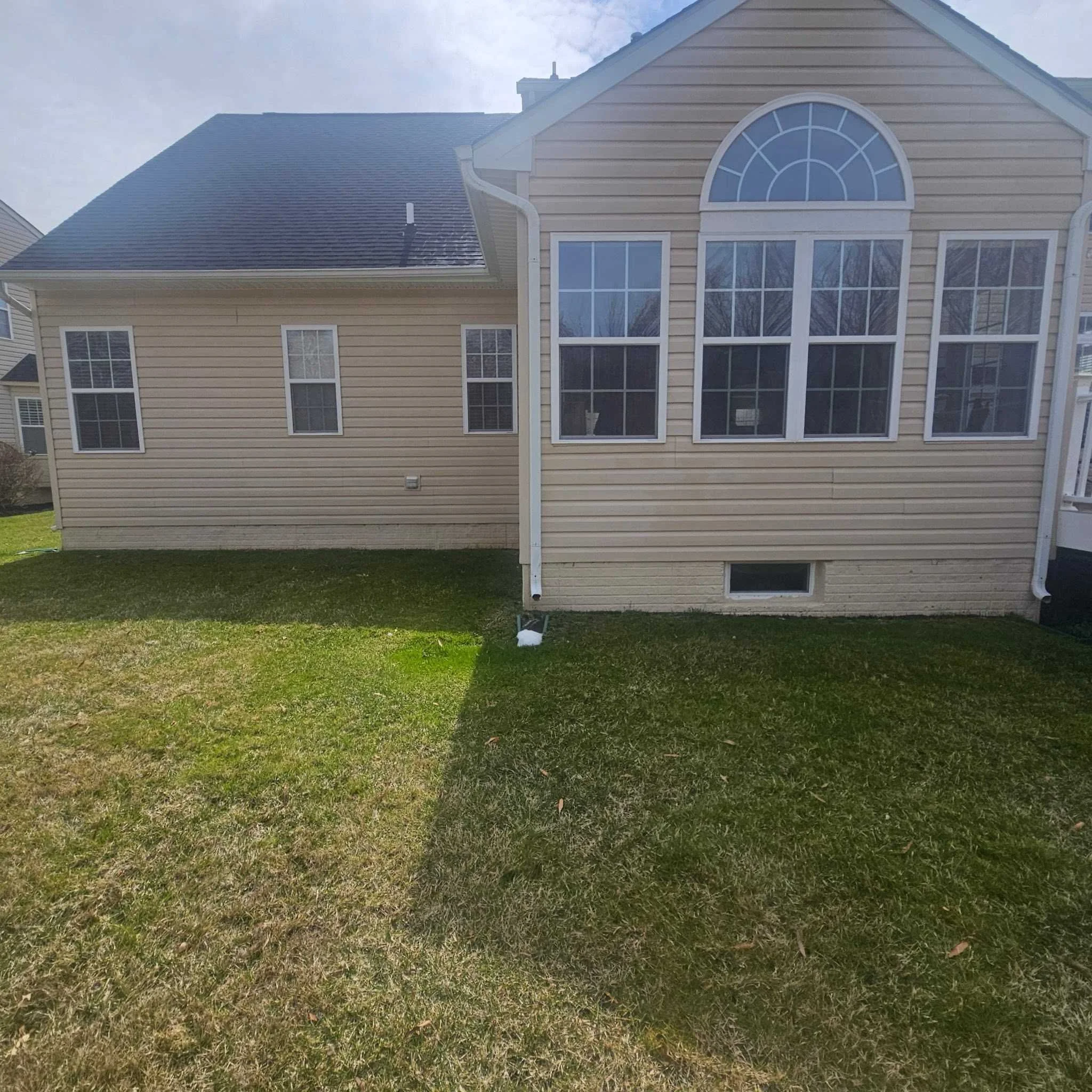 Side view of a beige house with multiple windows and a backyard with grass, partly shadowed.