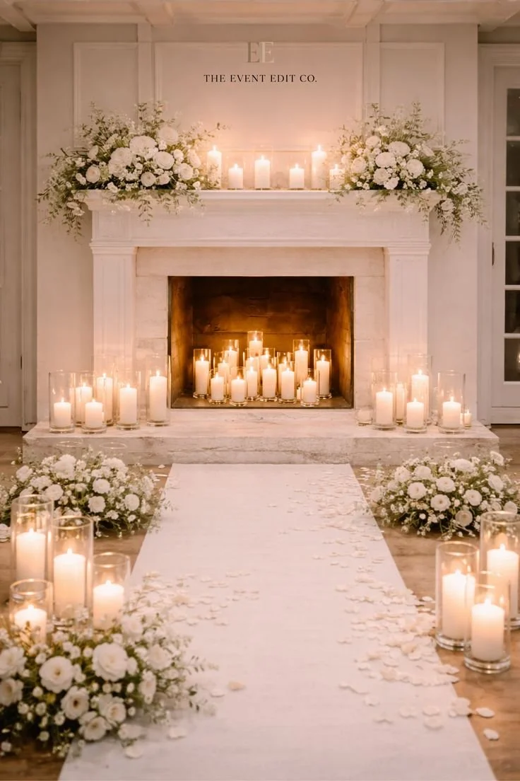 Elegant wedding ceremony setup with white floral arrangements, numerous candles, and a white aisle runner leading to a decorated fireplace.