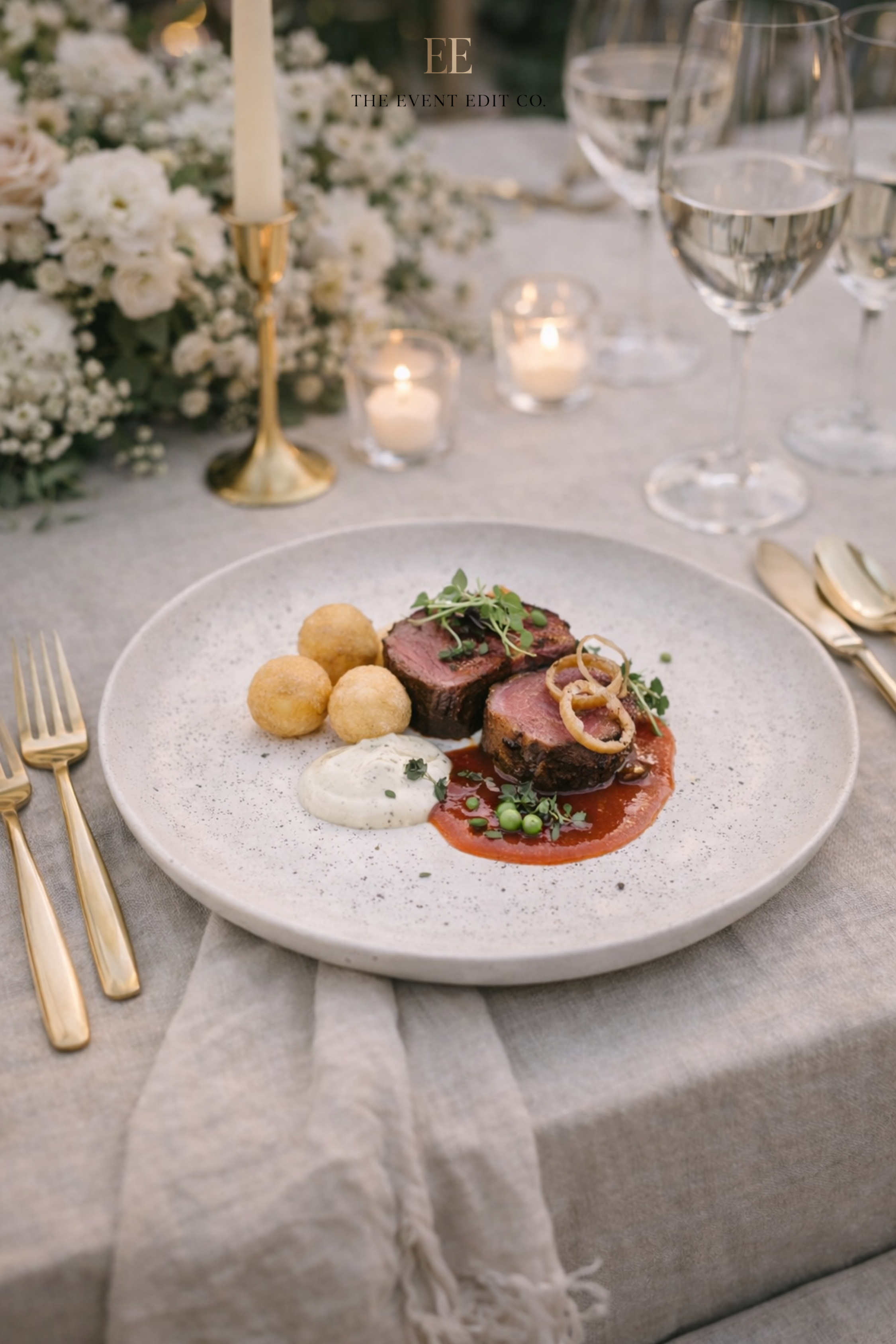 Elegant table setting featuring a white plate with sliced beef tenderloin topped with microgreens and caramelized onions, accompanied by fried croquettes and a dollop of white sauce, on a beige linen tablecloth, surrounded by wine glasses, candles, a