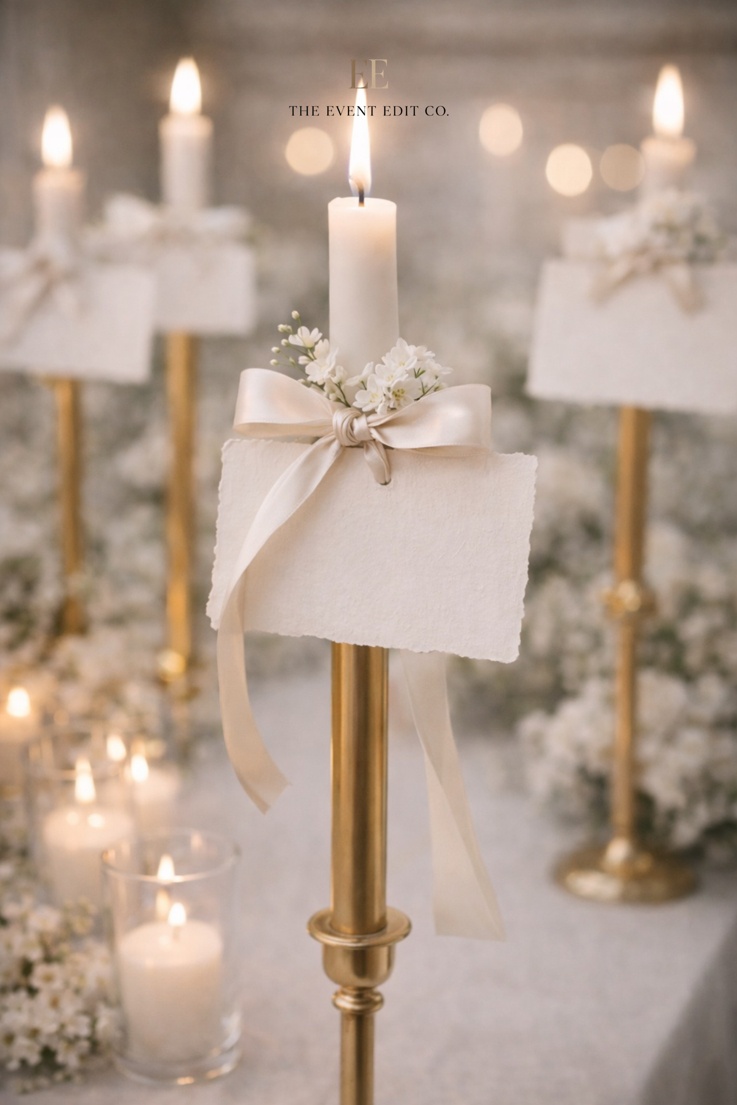 Elegant candle centerpiece with a lit white candle, tied with a satin ribbon and small white flowers, set on a gold stand at a formal event, with blurred candles and floral decorations in the background.