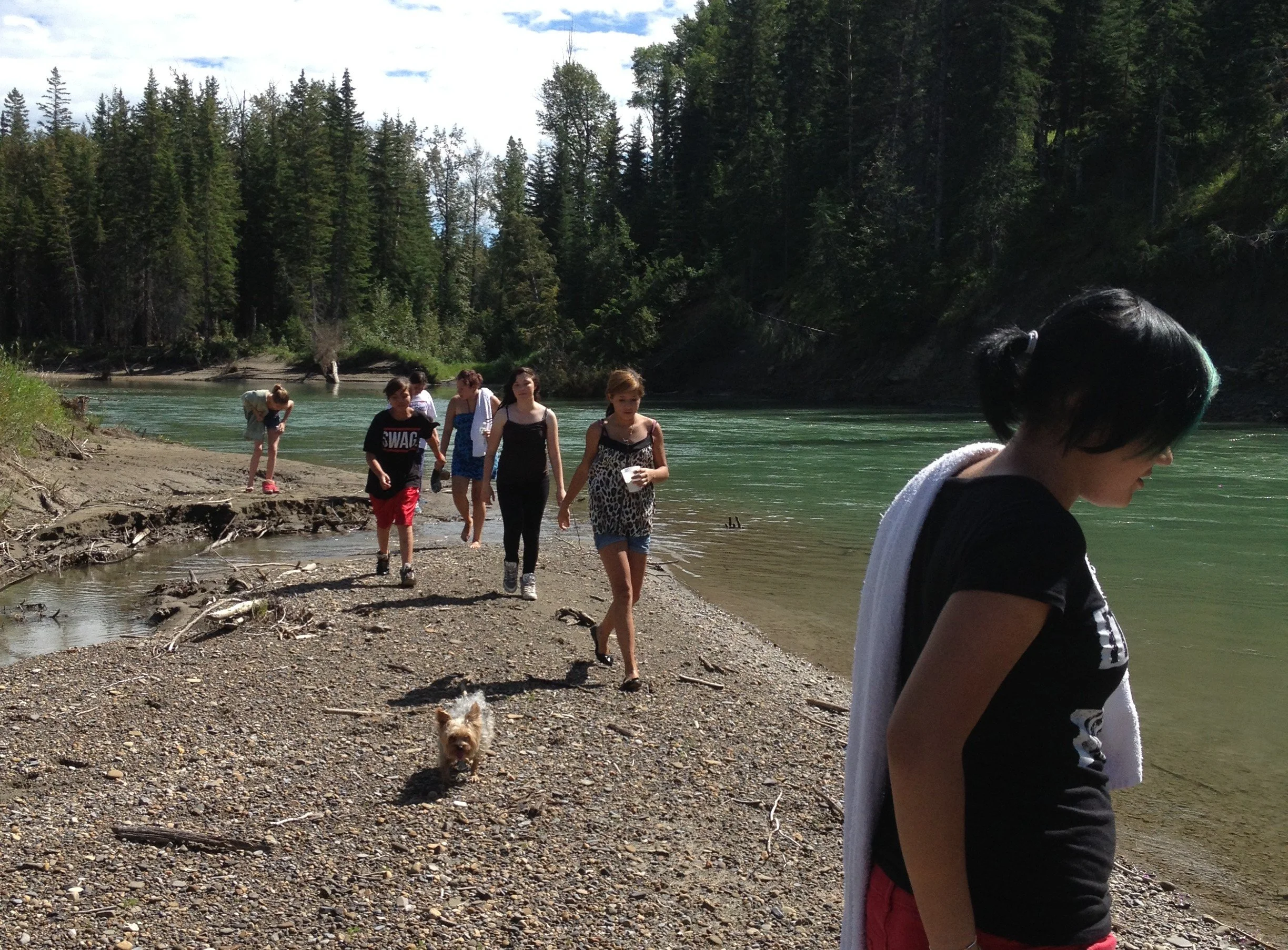 Group of people, including children and a woman, walking along a rocky riverside path with a small dog, surrounded by dense green forest under a partly cloudy sky.