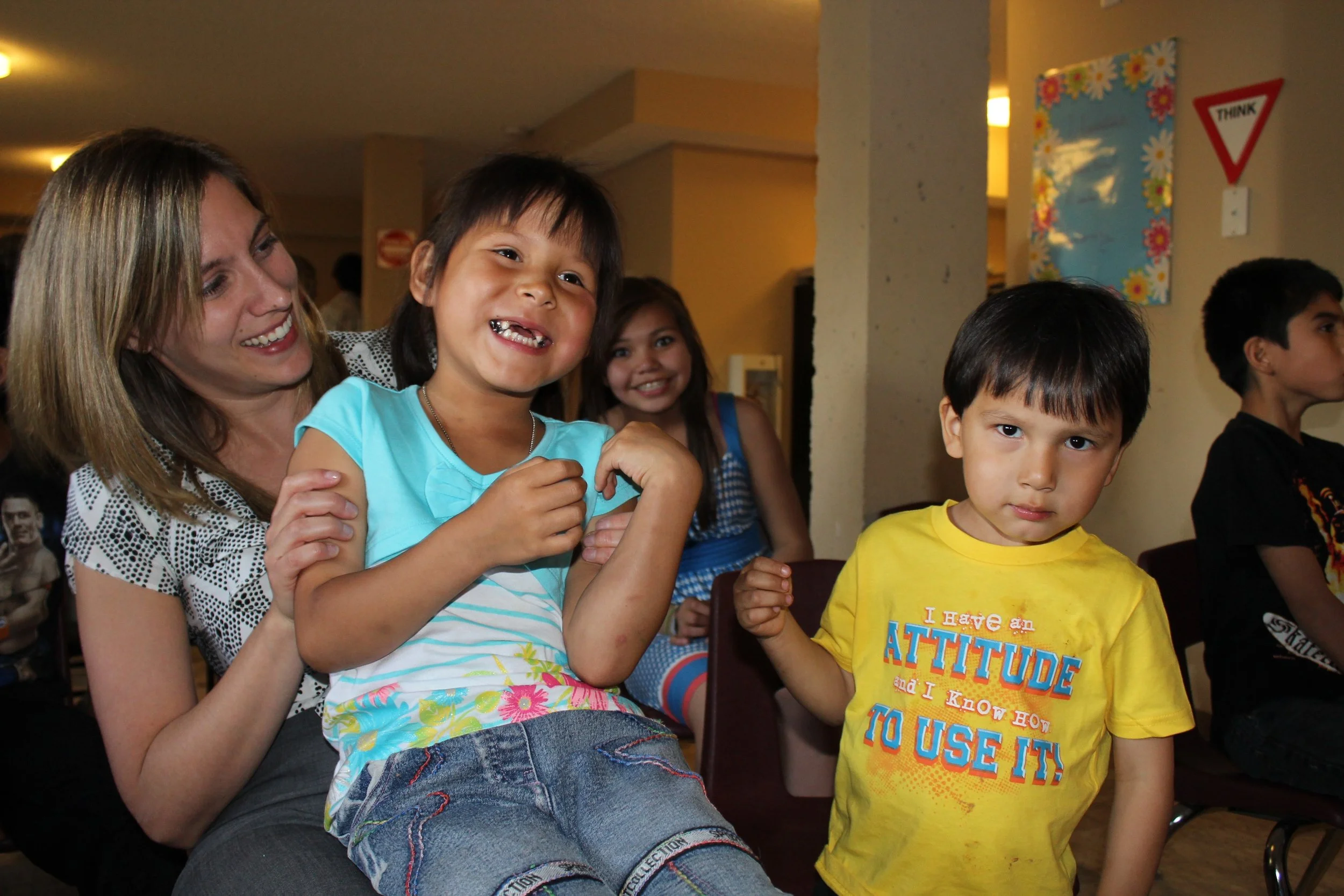 A smiling woman sitting with children, one girl with a bright smile wearing a blue shirt and denim shorts, and a boy in a yellow shirt with a serious expression, in a colorful indoor setting with other children in the background.