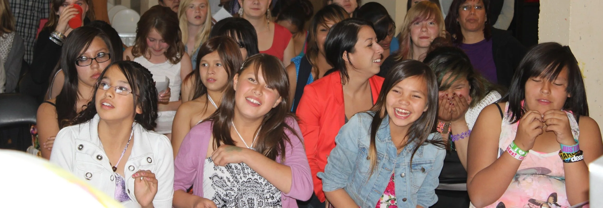 A group of children and a woman in an audience, many of them smiling and laughing, sitting close together at an event.