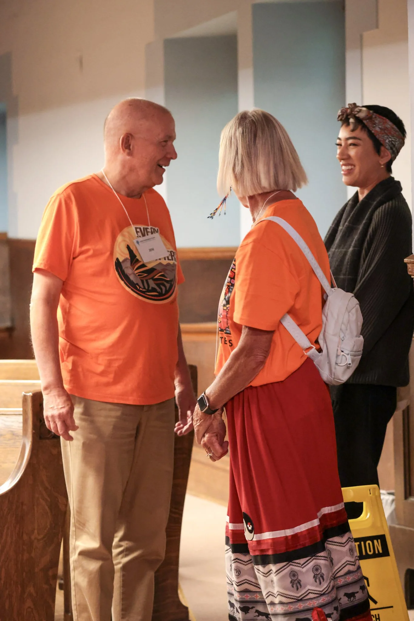 Three people, two women and one man, are smiling and talking in what appears to be a community or conference setting. The man and one woman are wearing matching orange shirts with a logo. The other woman has a white backpack and colorful earrings.