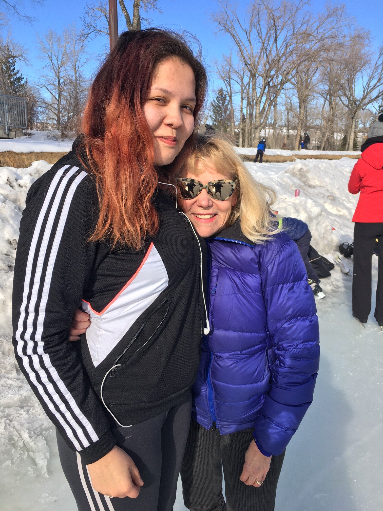 Two women posing outdoors in a snowy setting with trees, one with red hair wearing a black and white sports jacket, and the other with blonde hair, sunglasses, and a purple puffy jacket.