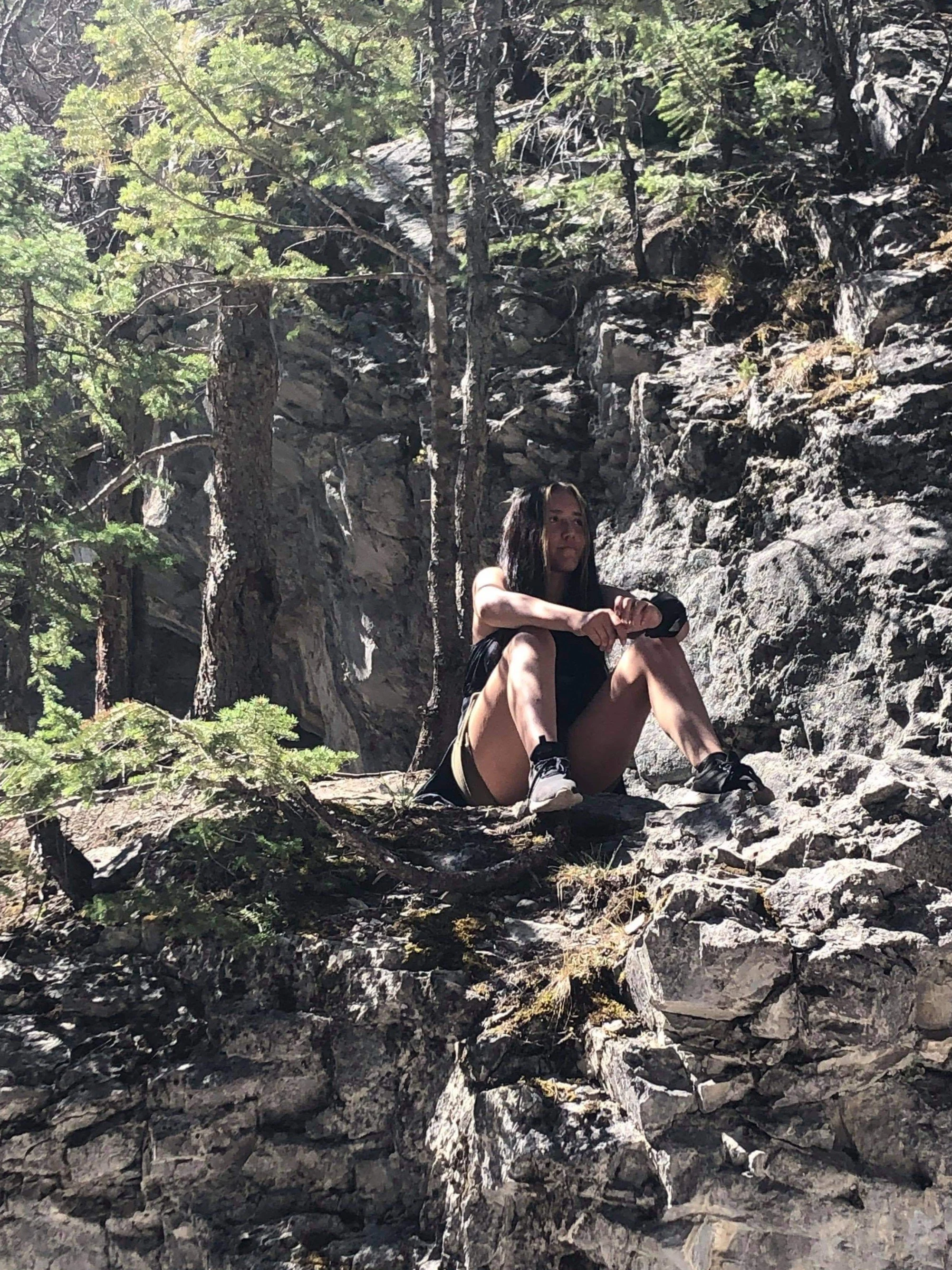A woman sitting on rocky terrain surrounded by trees in a forest, looking contemplative while resting.