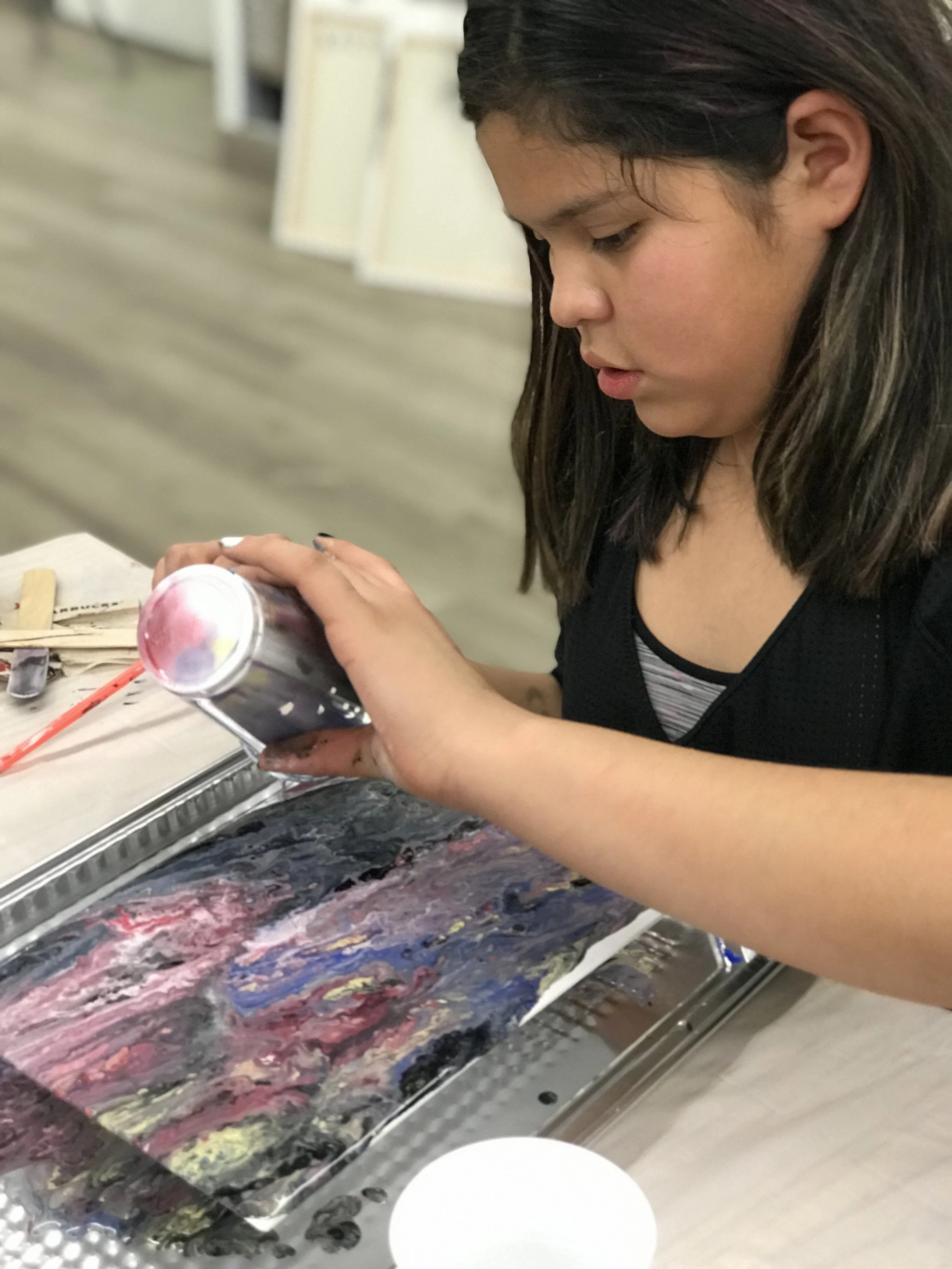 A girl with dark hair and a black shirt is pouring paint from a can onto a canvas, creating colorful abstract art.