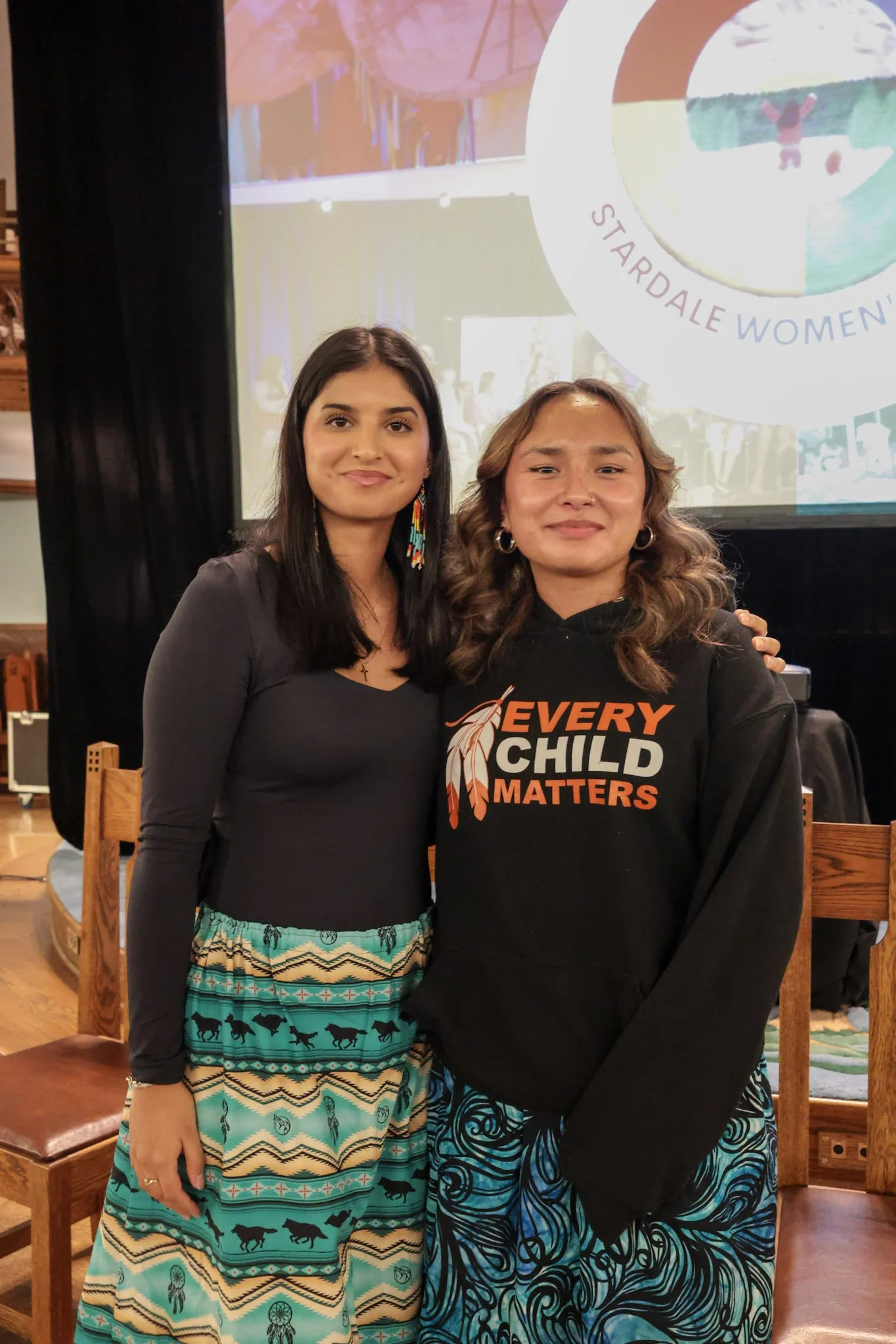 Two young women standing arms around one another, smiling.